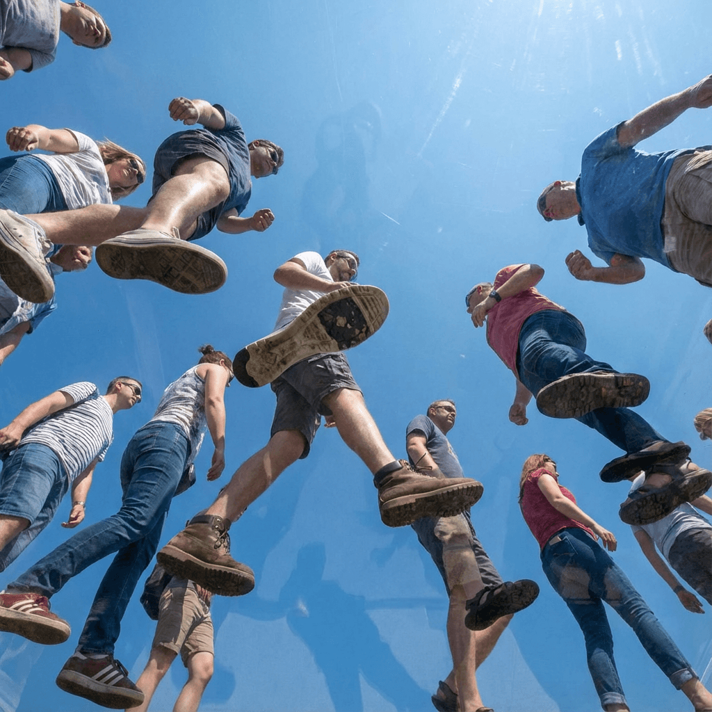 View from under a plane of completely transparent plexiglass. We are looking straight up. Many people are walking on the plexiglass. In the background is a pure blue sky. No buildings visible. No edges or separations in the plexiglass. It’s as if the plexiglass isn’t even there. We are very close to the people. The people are in the process of walking. The people are walking generally from left to right. The soles of their shoes are very close to the foreground.