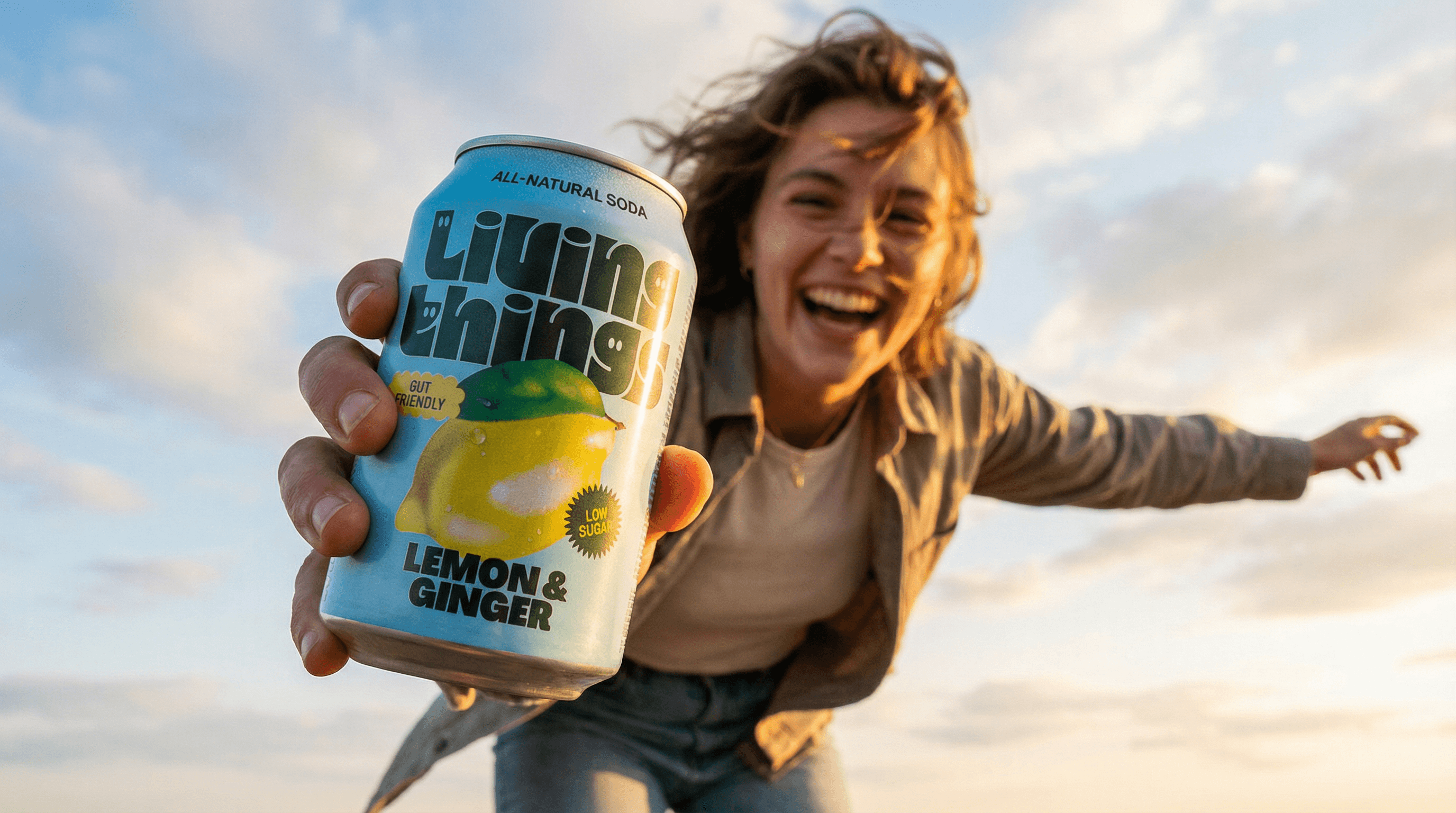 The photo captures a spontaneous, high-energy lifestyle moment with a strong sense of movement and joy.
A person is photographed from a low angle outdoors, arm extended toward the camera while holding a bright blue beverage can. The can dominates the foreground, enlarged by perspective, while the person’s face sits slightly behind it, smiling broadly and mid-laugh. The gesture feels playful and inviting, as if offering the drink to the viewer.
The background is an open sky with soft clouds, adding freedom and openness to the scene. Wind-tousled hair and casual styling enhance the feeling of motion and immediacy, like a moment grabbed on the fly rather than carefully staged.
Lighting appears natural and directional, likely late afternoon or early evening, creating gentle contrast and a cinematic feel. The saturated blue of the can pops strongly against the muted sky and darker clothing, making the product visually unmistakable without isolating it from the moment.
Overall, the image feels carefree, youthful, and authentic, positioning the product as part of an energetic, social experience rather than a posed advertisement.