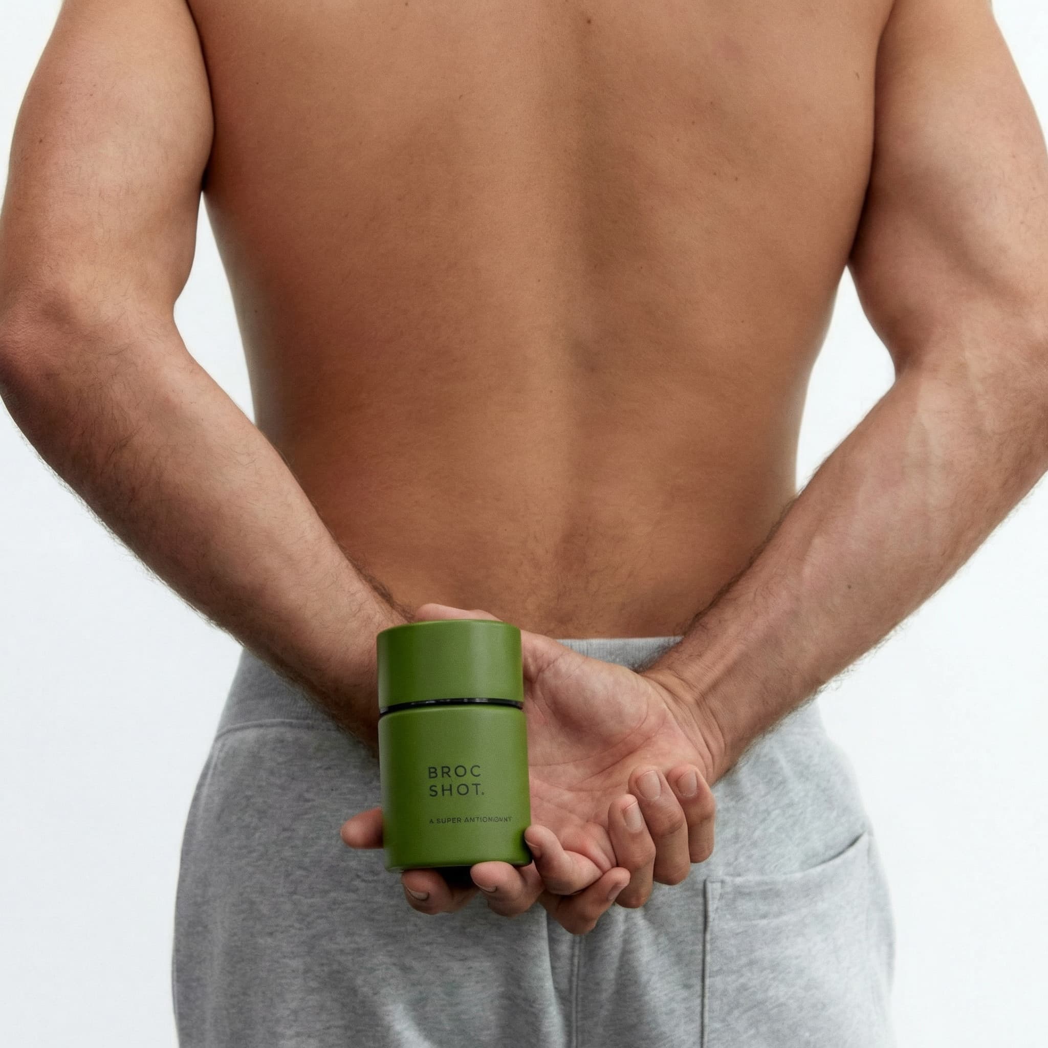 An intimate wellness product photograph showcasing masculine self-care. Shot from behind, a shirtless male torso is centered in the frame, skin tones warm and natural. The subject wears light gray sweatpants, hands clasped behind the back holding a dark green glass supplement bottle. The composition is minimalist and clean, shot against a pure white seamless background. The lighting is soft and diffused, creating gentle shadows that define the body's contours. The mood is calm, modern, and health-focused—emphasizing natural wellness and mindful masculinity. The color palette is muted and earthy: warm skin tones, soft gray fabric, deep forest green glass. The aesthetic is editorial and refined, balancing intimacy with professionalism. Focus is sharp on the central figure with the bottle as a subtle focal point.
