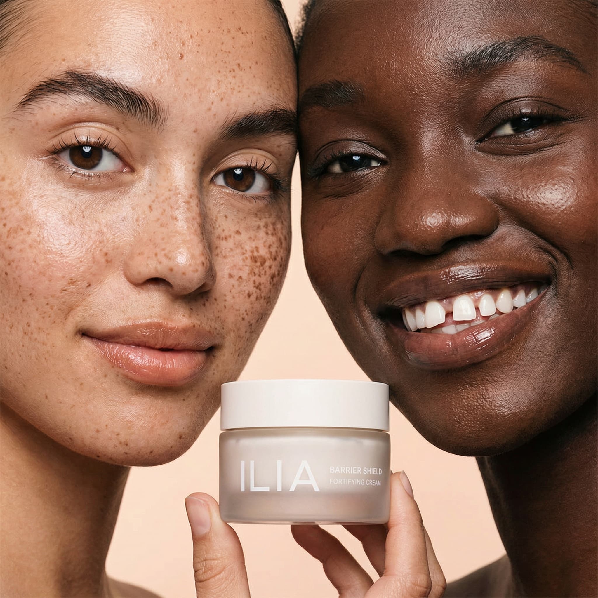 A striking beauty editorial celebrating diversity and natural skin. Two faces fill the frame from opposite sides—on the left, a fair-skinned person with freckles and warm brown eyes; on the right, a dark-skinned person with a radiant smile showing white teeth. Both have glowing, unretouched skin that celebrates natural beauty marks and texture. Between them, held center-frame, is a frosted glass jar of ILIA 'BARRIER SHIELD FORTIFYING CREAM'—the hero product. The composition is intimate and bold, shot against a soft peachy-beige gradient background. Lighting is even and flattering, highlighting the warmth and natural radiance of both skin tones. The mood is joyful, inclusive, and empowering—showcasing beauty in all its forms. The color palette is warm and earthy: freckled beige, rich brown skin, peachy background, and the creamy product jar. The aesthetic is modern, clean, and authentic—editorial quality with genuine human connection. Focus is crisp on both faces and the product label.