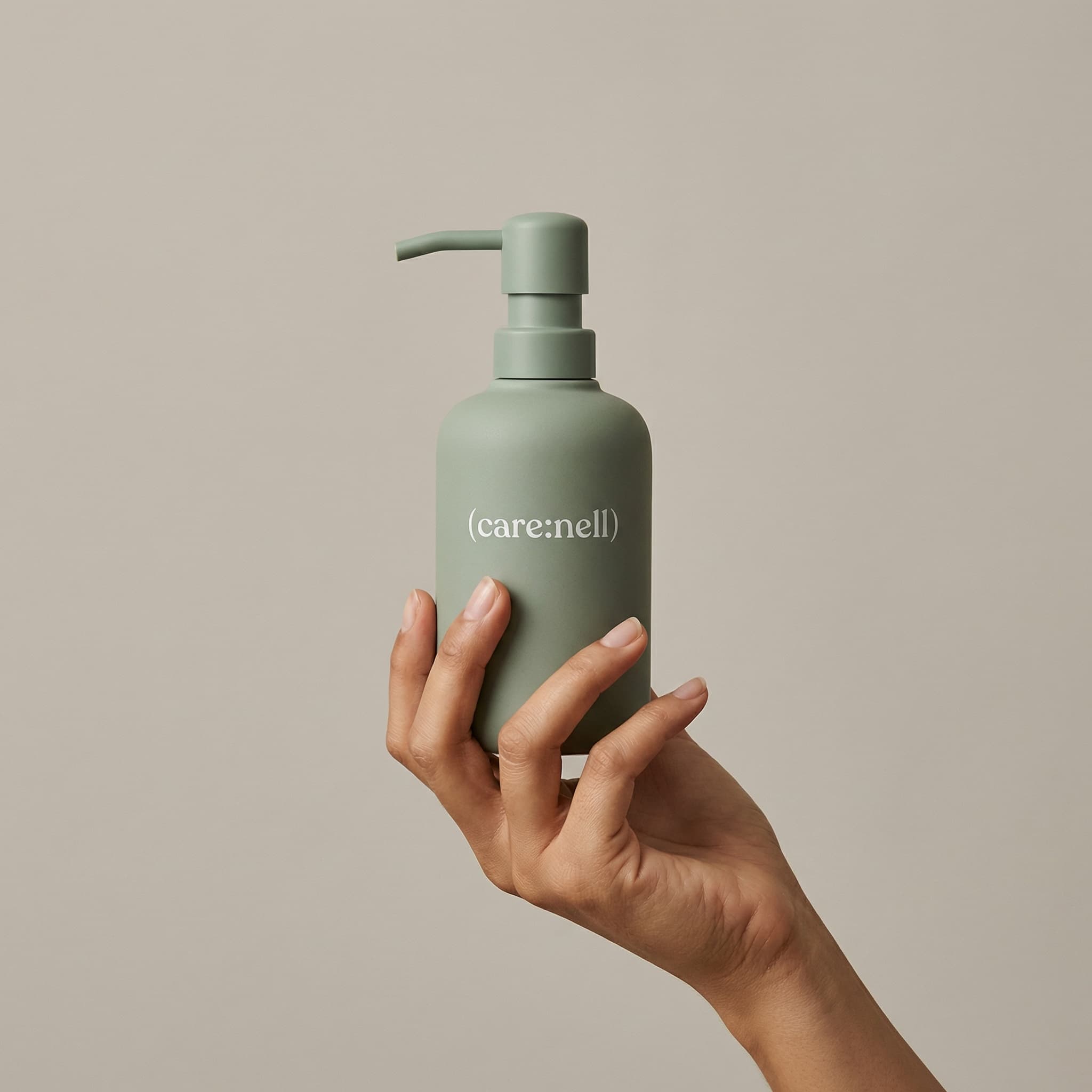 A minimalist wellness product shot with organic simplicity. A hand enters from the bottom-right, fingers delicately cradling a matte sage-green bottle with a pump dispenser. The bottle has clean typography reading '(care:nell)' in white. The composition is centered and balanced, shot against a soft neutral beige-gray seamless background. The lighting is gentle and diffused, creating subtle shadows that add depth without harshness. The hand is natural and unmanicured, with warm skin tones that contrast beautifully against the cool sage green. The mood is calm, sustainable, and thoughtfully designed—evoking mindful consumption and eco-conscious beauty. The color palette is earthy and muted: soft eucalyptus green, warm beige background, natural skin. The aesthetic is refined minimalism—clean, modern, and effortlessly sophisticated. Focus is sharp on the product label and the hand holding it. The overall feeling is serene and grounded.
