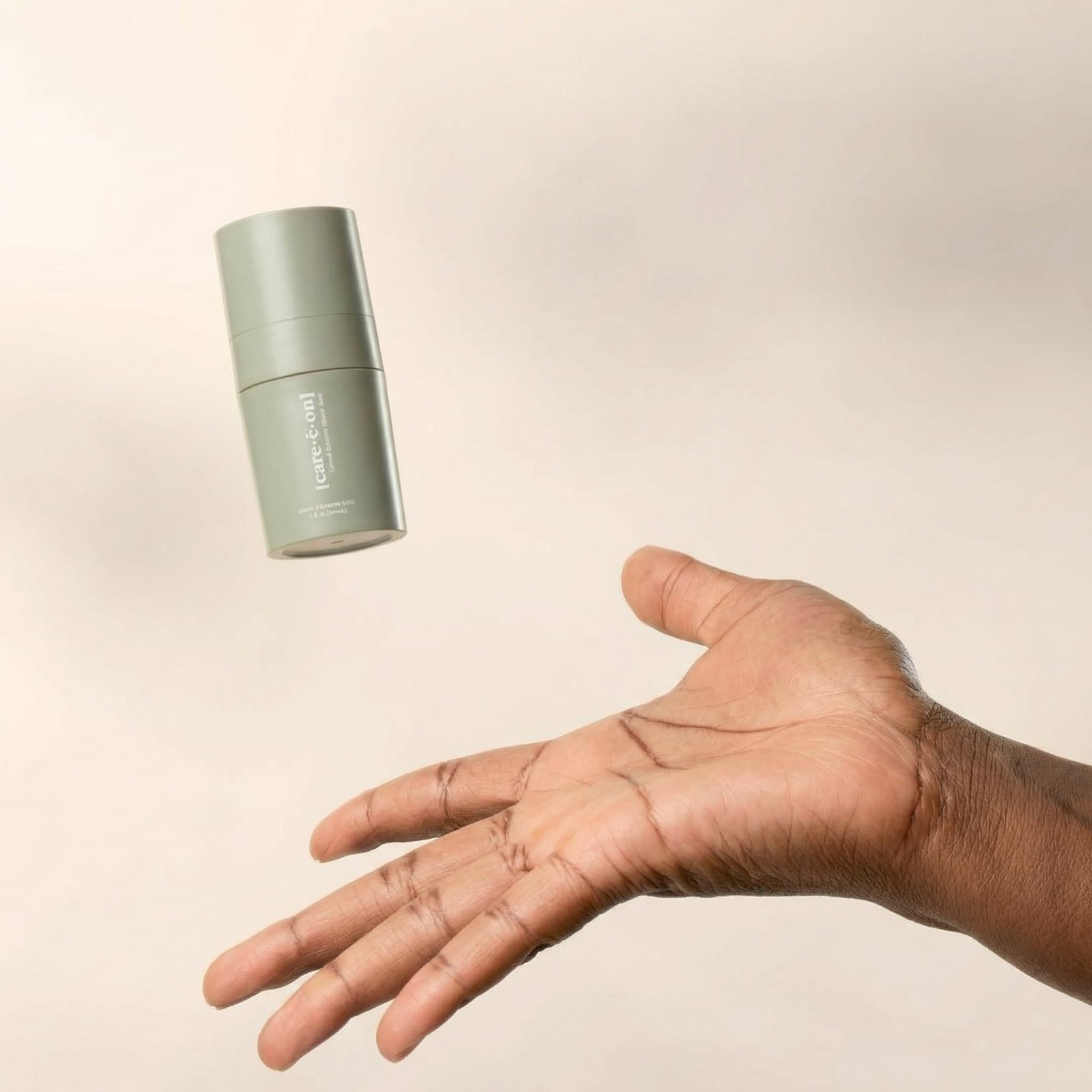 A playful beauty product photograph capturing spontaneous motion. A matte sage-green cylindrical pump bottle floats mid-air in the upper-left, angled slightly as if just released. Below and to the right, an open hand with warm brown skin reaches upward, palm facing up, fingers naturally spread—as if catching or about to receive the product. The composition is dynamic yet balanced, shot against a soft gradient background that transitions from warm cream to pale beige. The lighting is gentle and diffused, creating subtle shadows that add dimensionality. The mood is joyful, carefree, and confident—evoking trust and effortless beauty. The bottle reads '(care:nell)' in white typography. The color palette is warm and organic: soft eucalyptus green, natural brown skin, peachy-cream background. The aesthetic is modern and aspirational with a playful twist—editorial quality meets authentic human energy. Focus is sharp on both the floating bottle and the extended hand.