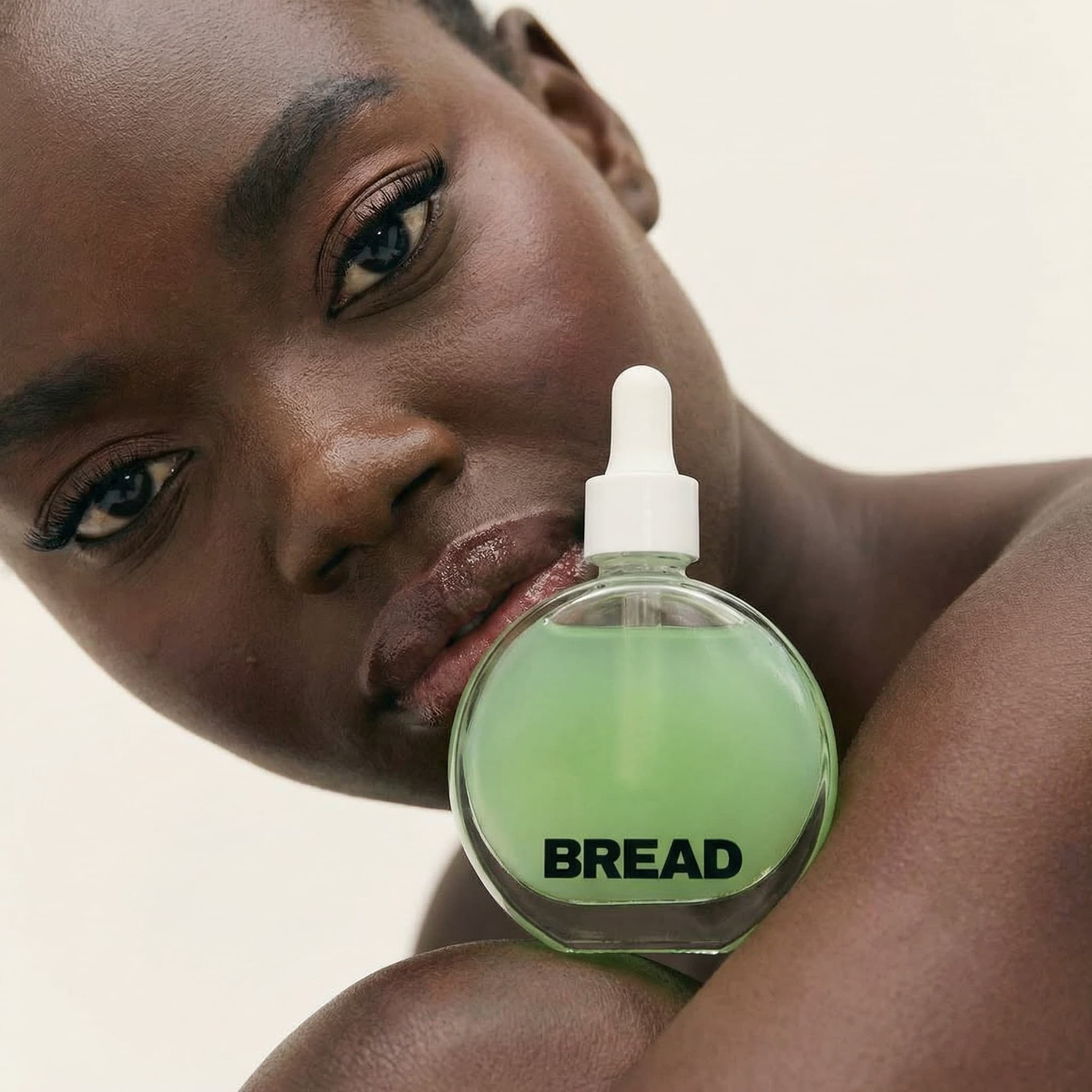 An intimate and artistic beauty editorial shot from a low angle. A Black model reclines, their face tilted diagonally across the frame with a serene, contemplative expression. Their skin is radiant with a natural glow, eyes striking with defined brows and lashes. In the foreground center, resting against their shoulder, is a spherical glass bottle filled with translucent pale green serum—labeled 'BREAD' in bold black typography. The bottle has a white dropper cap. The composition creates a powerful connection between face and product, with smooth dark skin contrasting beautifully against the luminous green liquid. Shot against a soft off-white seamless background with gentle, flattering lighting that highlights skin texture and the product's transparency. The mood is confident, intimate, and aspirational—celebrating natural beauty and self-care. The color palette is warm and organic: rich brown skin, soft mint-green serum, neutral cream background. The aesthetic is modern editorial with artistic sophistication. Focus is sharp on both the face and product bottle.
