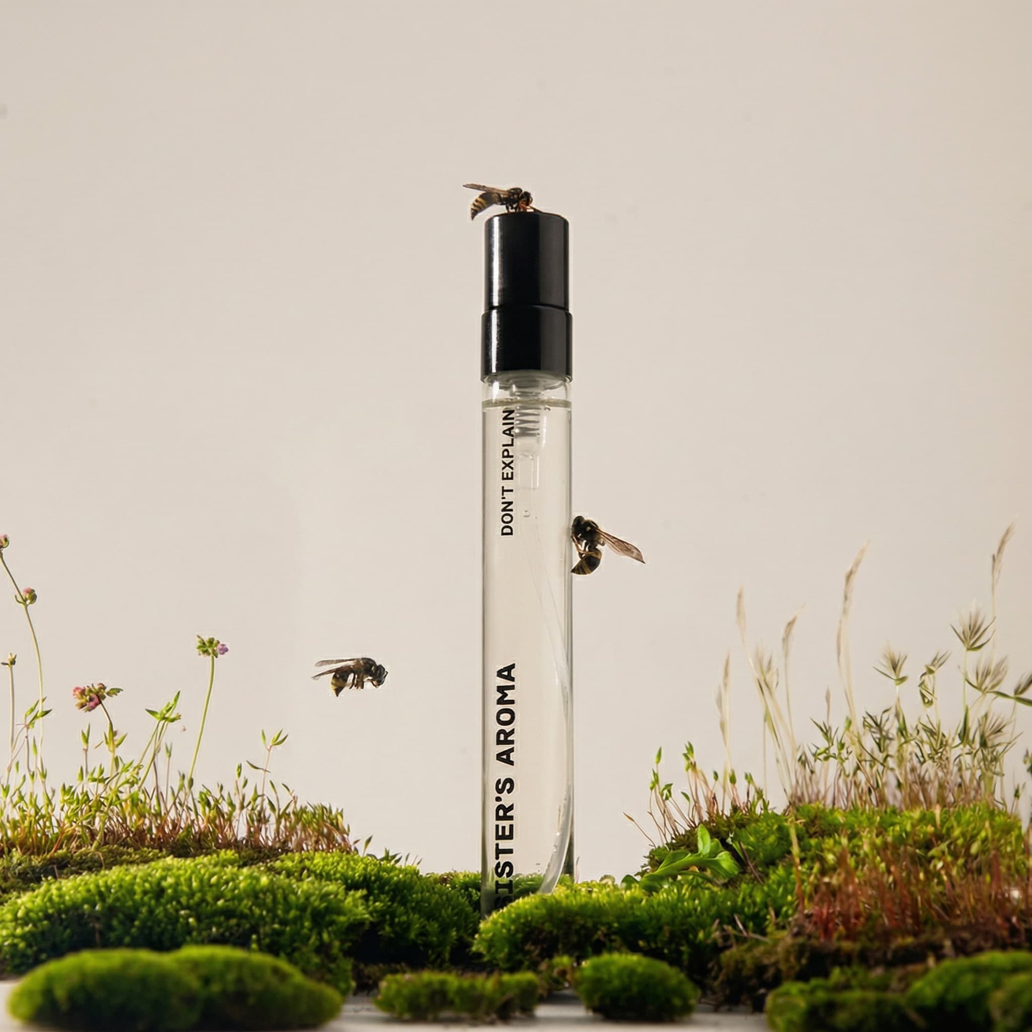 A whimsical and naturalistic beauty product photograph blending macro nature with minimalism. A tall, slender clear glass serum bottle with a matte black cap stands vertically center-frame, rising from a lush miniature landscape. The foreground is alive with vibrant green moss mounds, delicate wildflowers on thin stems, and wispy dried grasses creating a fairy-tale ecosystem. Three bees hover mid-flight around the bottle—one perched on the cap, others buzzing nearby—adding organic movement and life. The bottle is labeled 'DON'T EXPLAIN' and 'SISTER'S AROMA' in black vertical typography. The composition is surreal yet grounded, shot against a soft neutral beige background with gentle natural lighting that makes the moss glow. The mood is enchanting, eco-conscious, and poetic—celebrating nature-inspired beauty and sustainability. The color palette is earthy and fresh: rich emerald moss, warm honey-toned bees, clear glass, matte black, soft beige sky. The aesthetic is editorial with a touch of magical realism—like stepping into a miniature world. Focus is balanced between the bottle and the living landscape.
