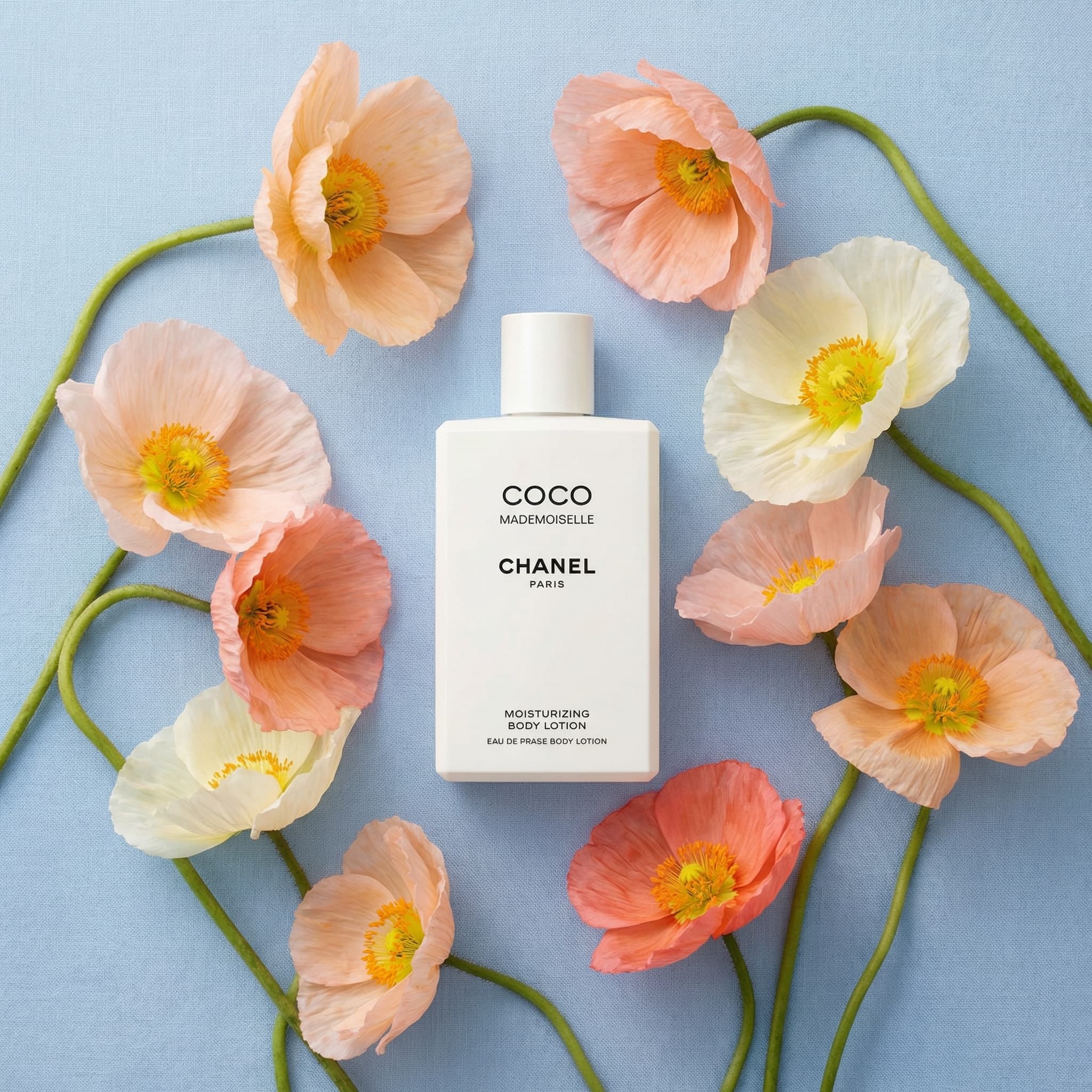 An elegant flatlay product photograph with botanical artistry. A pristine white CHANEL 'COCO MADEMOISELLE' moisturizing body lotion bottle lies centered against a soft powder-blue textured background. Surrounding it in a graceful arrangement are delicate poppy flowers in romantic shades—soft peach, blush pink, creamy white, and coral—with papery translucent petals and vibrant orange stamens. The poppies have elegant curved green stems that create organic lines flowing around the product. The composition is balanced and poetic, shot from directly above with even, diffused lighting that highlights the delicate petal texture and the clean white bottle. The mood is luxurious, feminine, and spring-like—evoking French elegance and natural beauty. The color palette is soft and romantic: powder blue, peachy coral, blush pink, creamy white, fresh green stems, golden stamens. The aesthetic is high-end editorial—refined, sophisticated, and effortlessly beautiful. Focus is crisp on both the Chanel branding and the flower details.