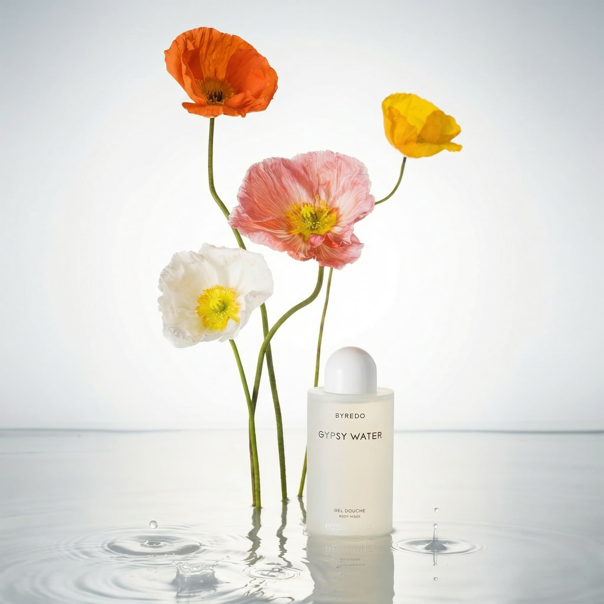 A serene and artistic product photograph merging beauty with nature. Four delicate poppy flowers on long green stems stand gracefully in shallow, reflective water—vibrant orange, sunny yellow, soft coral-pink, and pure white, each with bright yellow stamens and papery translucent petals. The flowers rise from the water surface, creating gentle ripples and mirror reflections. Beside them stands a pristine white BYREDO 'GYPSY WATER' gel douche bottle with a rounded dome cap, also reflected in the water. The composition is ethereal and balanced, shot against a soft gradient background that transitions from pale gray to white. Lighting is clean and diffused, highlighting the delicate petal texture and the water's crystalline clarity. The mood is tranquil, luxurious, and poetic—evoking spa-like serenity and natural beauty. The color palette is fresh and vibrant: bright orange, golden yellow, blush pink, creamy white, fresh green stems, clear water. The aesthetic is high-end editorial with an artistic, zen-like quality. Focus is sharp on the flowers and product, with beautiful bokeh reflections in the water.
