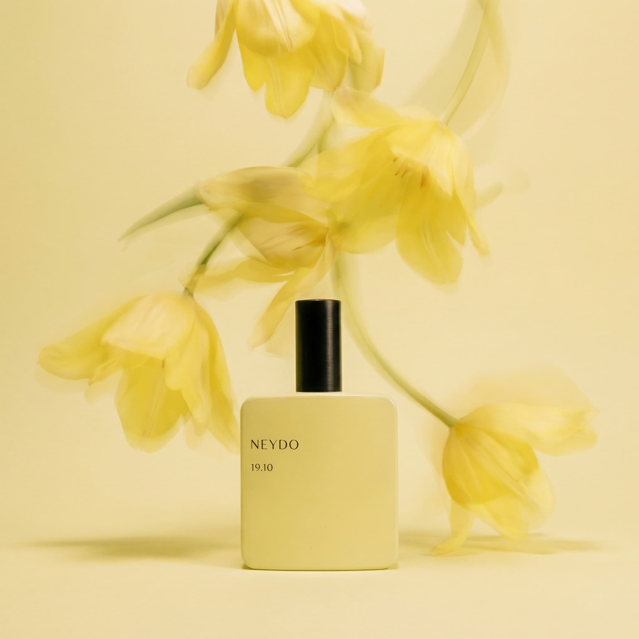 An ethereal and artistic fragrance photograph with dreamy motion. A soft butter-yellow NEYDO '19.10' perfume bottle with a matte black cap sits centered in the lower half of the frame against a matching pale yellow seamless background. Above and behind it, delicate yellow flower petals—possibly tulips or irises—are captured in motion blur, creating ghostly, flowing shapes that dance through the frame. The motion creates a sense of movement frozen in time, with soft layered forms and translucent overlapping petals. The composition is minimalist yet dynamic, shot with a shallow depth of field that keeps the bottle crisp while the background blooms into abstract yellow shapes. Lighting is soft and even, creating a gentle glow throughout. The mood is artistic, elegant, and contemplative—evoking fleeting beauty and sensory experience. The color palette is monochromatic yellow: butter cream, soft lemon, golden wheat, with a black accent from the cap. The aesthetic is fine art photography meets high-end perfume editorial—poetic, refined, and otherworldly. Focus is sharp on the bottle with intentional motion blur on the flowers.
