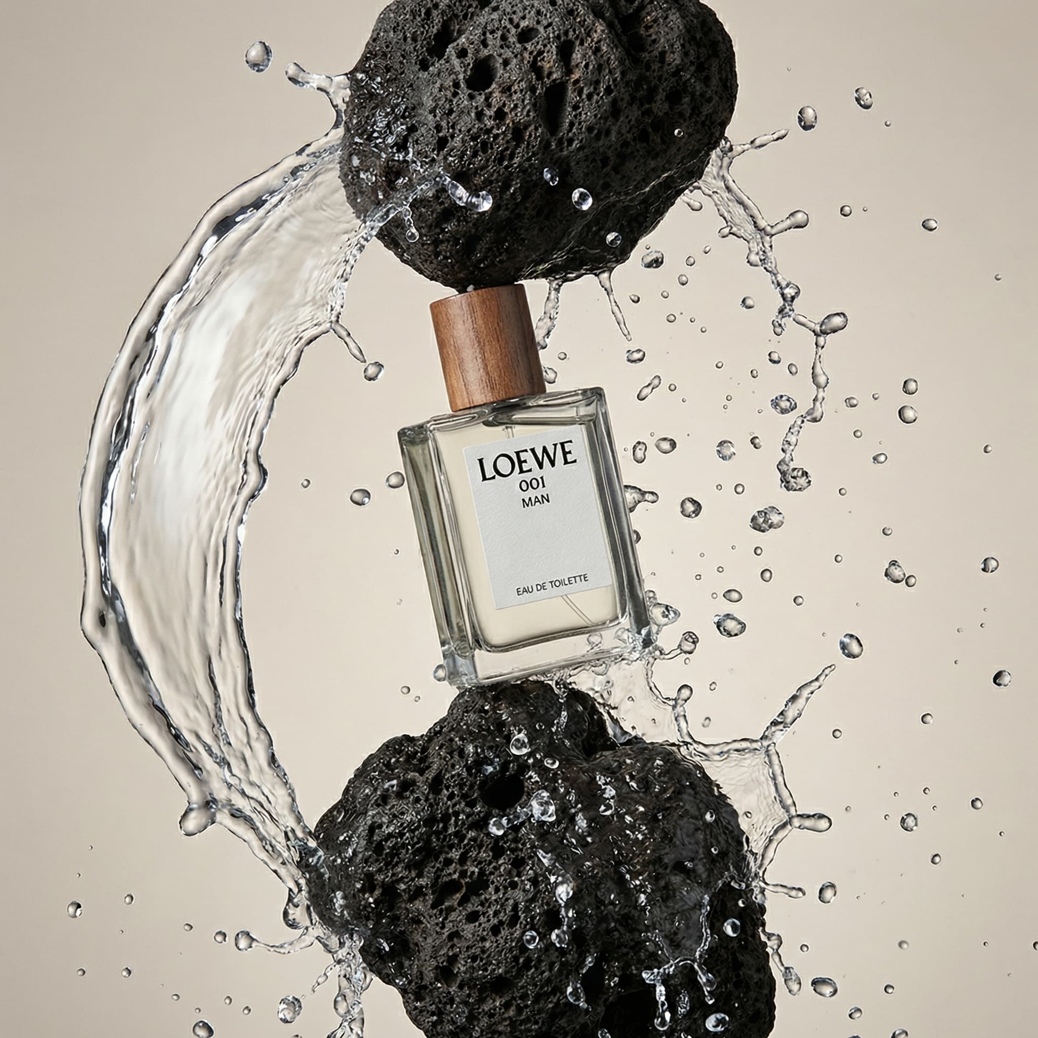 A dynamic and surreal fragrance photograph capturing motion and nature. A square glass LOEWE '001 MAN' eau de toilette bottle with a warm wood-toned cap is suspended center-frame, balanced precariously between two dark volcanic rocks—one above, one below—as if defying gravity. Water splashes dramatically around the bottle in frozen motion, with arcing streams on the left and cascading droplets scattered throughout the composition. Hundreds of tiny water droplets float in the air, catching the light like crystal beads. The rocks are textured and porous, contrasting beautifully with the sleek glass bottle. Shot against a soft neutral beige background with studio lighting that highlights the water's transparency and creates a sense of arrested movement. The mood is bold, sculptural, and elemental—evoking the raw power of nature meeting refined luxury. The color palette is earthy and organic: dark charcoal rock, warm wood cap, clear glass with pale liquid, transparent water, soft beige background. The aesthetic is high-concept editorial with a surrealist edge—part fine art, part product photography. Focus is sharp on the bottle and rocks with beautiful detail in the frozen water splash.