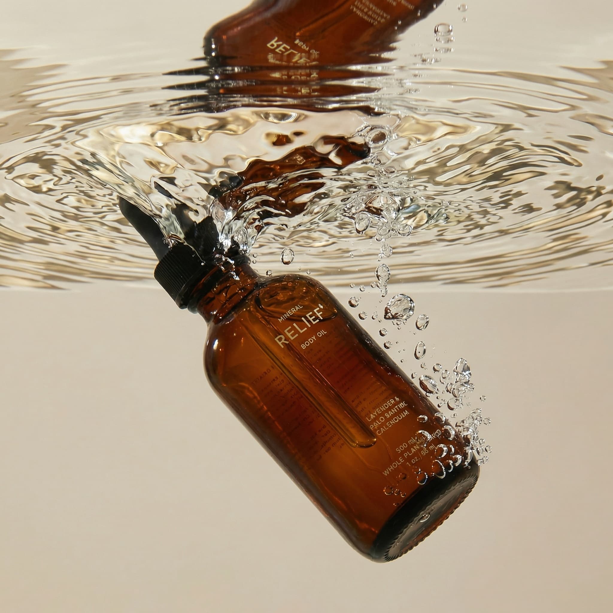A stunning underwater product photograph with liquid refraction. A deep amber glass bottle of RELIEF 'MINERAL BODY OIL' is submerged just below the water's surface, tilted at an angle with bubbles streaming upward. The bottle has elegant gold typography reading 'RELIEF' and 'BODY OIL' with additional text about lavender, palmarosa, and calendula. The water surface above creates mesmerizing ripples and refraction patterns—wavy distortions that bend light into abstract shapes. Below the waterline, the bottle is clear and crisp, while above the surface the reflections create a mirrored, dreamlike effect. Tiny bubbles cling to the bottle and float upward. The composition is split between underwater and surface views, shot against a soft neutral beige background visible through the water. Lighting is bright and clean, highlighting the transparency of the water and the rich amber color of the bottle. The mood is refreshing, spa-like, and luxurious—evoking hydrotherapy and natural wellness. The color palette is warm and organic: deep amber glass, gold text, crystal-clear water, soft beige background. The aesthetic is high-end editorial with a surreal, meditative quality. Focus is sharp on the submerged bottle with beautiful water distortion effects.