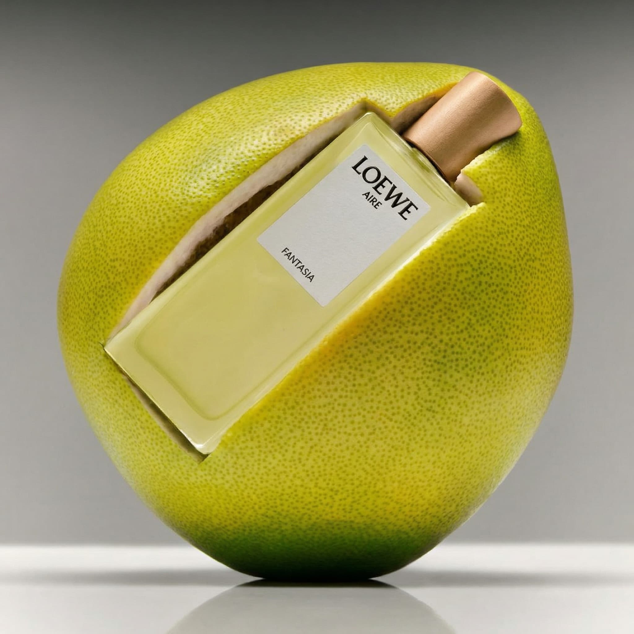 A playful and sculptural fragrance product photograph with natural elements. A pale yellow LOEWE 'AURA' perfume bottle with a rose-gold metallic cap rests nestled inside a hollowed-out pomelo or grapefruit. The citrus fruit is massive and spherical, with a vibrant chartreuse-yellow textured peel that gradients to deeper green at the bottom. The bottle fits perfectly in the carved-out cavity, creating a beautiful pairing of product and ingredient. The composition is shot from a slight overhead angle against a neutral gray gradient background, with the fruit casting a soft shadow on a reflective surface below. Lighting is clean and studio-perfect, highlighting the fruit's dimpled texture and the bottle's elegant simplicity. The mood is fresh, citrus-forward, and luxurious—directly connecting the fragrance to its natural inspiration. The color palette is vibrant and cohesive: bright chartreuse peel, pale yellow bottle, rose-gold cap, soft gray background. The aesthetic is high-end editorial with a conceptual twist—playful yet sophisticated, nature meets luxury. Focus is sharp on both the fruit texture and the bottle label.