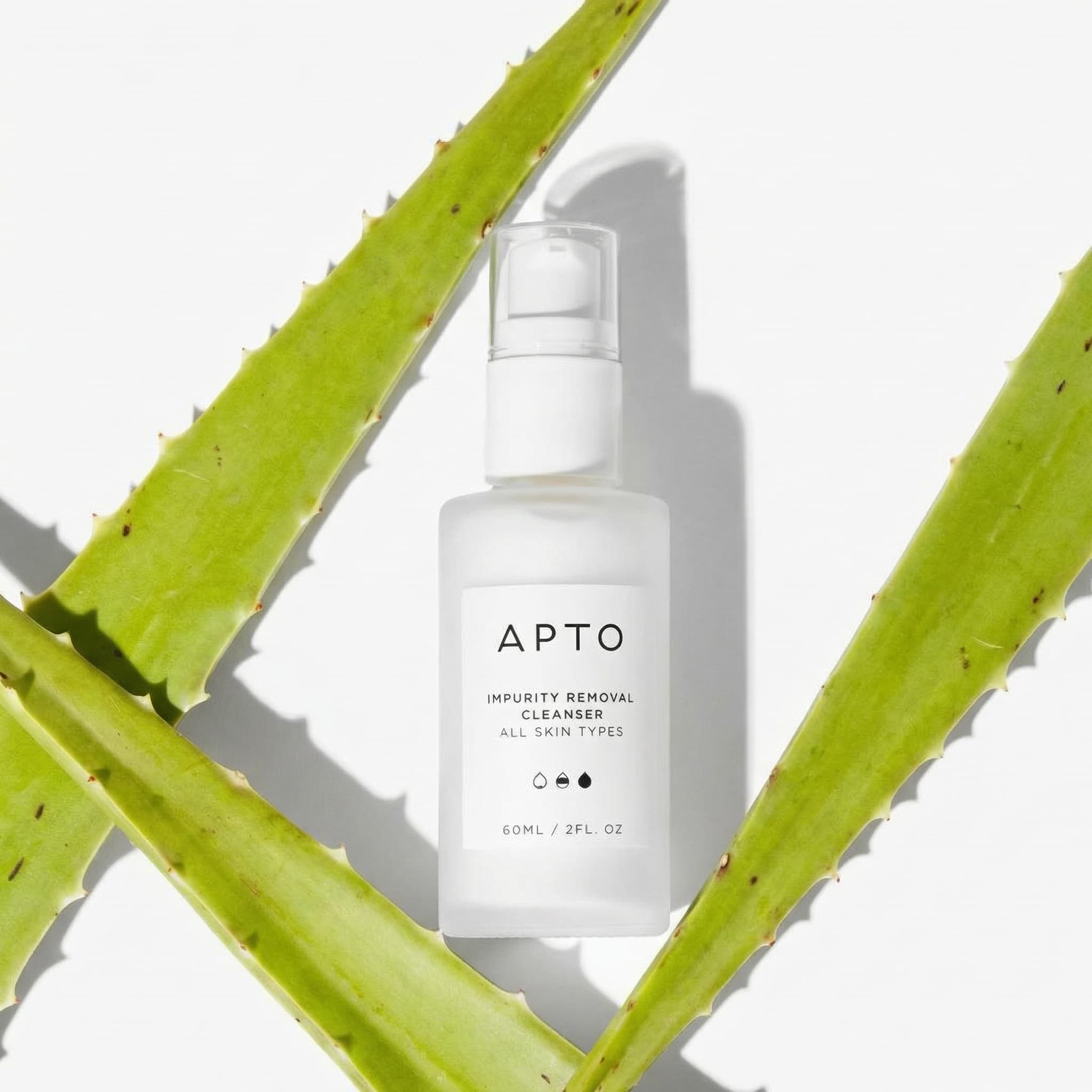 A clean and minimalist beauty product photograph with botanical framing. A frosted white glass APTO 'IMPURITY REMOVAL CLEANSER' bottle with a pump dispenser sits centered in the frame, casting a soft shadow on a pure white background. The bottle features minimal black typography reading 'APTO', 'IMPURITY REMOVAL CLEANSER', 'ALL SKIN TYPES', and '60ML / 2FL OZ' with simple iconography. Framing the bottle diagonally from both sides are two fresh aloe vera leaves with their characteristic serrated edges and spiky teeth visible along the sides. The leaves create natural leading lines that draw the eye to the product. The composition is architectural and balanced, shot with bright, even lighting that highlights the bottle's frosted texture and casts subtle shadows. The mood is fresh, pure, and clinically clean—evoking natural skincare and botanical ingredients. The color palette is crisp and natural: frosted white bottle, bright lime-green aloe, pure white background with soft gray shadows. The aesthetic is clean beauty minimalism—simple, honest, and ingredient-focused. Focus is sharp on the bottle label with the aloe leaves slightly softer but still detailed.