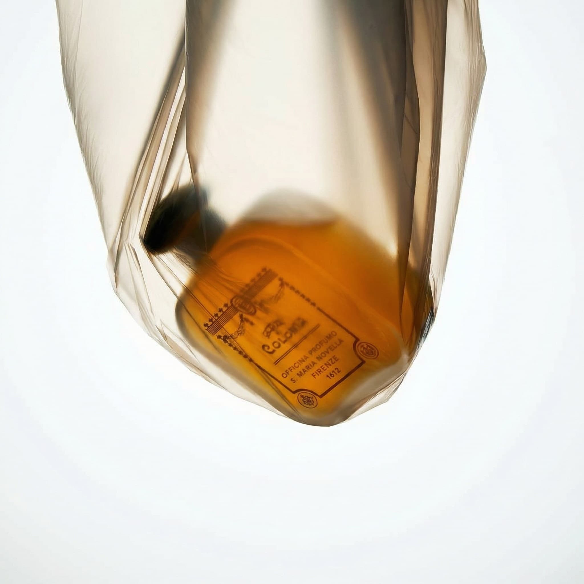 A luxury fragrance bottle with amber-colored liquid sealed inside a clear translucent plastic bag, suspended against a clean white background. The bottle is positioned upside down, creating a sense of weightlessness and ethereal suspension. Warm amber tones from the liquid contrast with the crisp white background. Minimalist editorial product photography with soft diffused studio lighting.