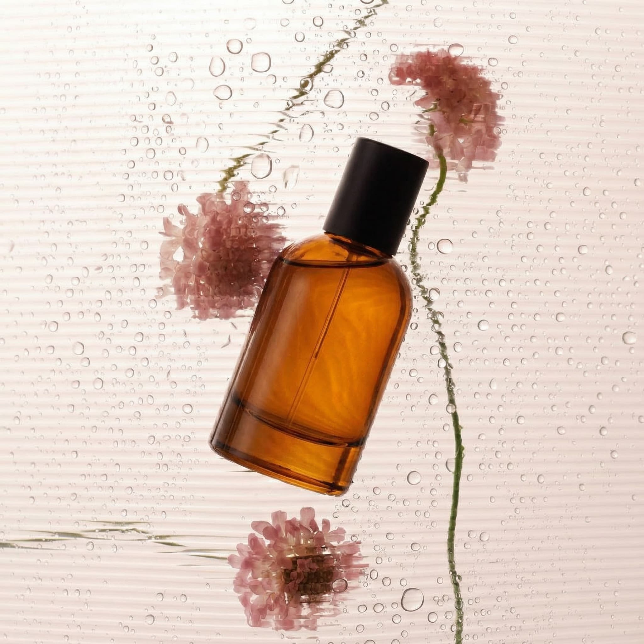 An amber glass fragrance bottle with a dark cap photographed behind textured water-droplet-covered glass, with delicate soft pink flowers floating and scattered around the bottle. Dreamy soft-focus botanical aesthetic with romantic natural diffused lighting. Muted pink and warm amber tones. High-end beauty product photography, delicate and feminine mood.