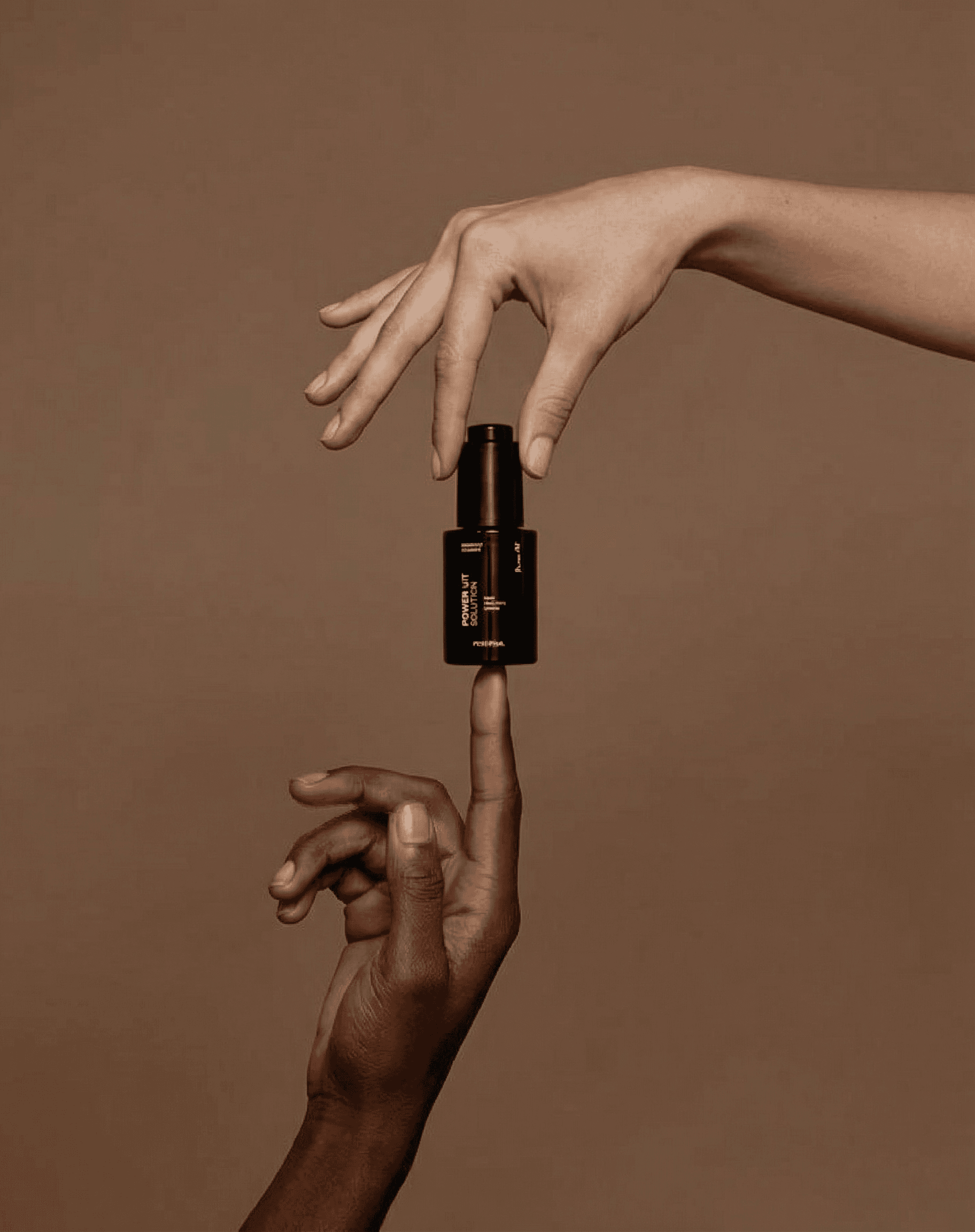 Two hands of different skin tones reaching toward each other, one lighter hand descending from above delicately pinching the top of a small dark cylindrical beauty bottle, one deeper-toned hand rising from below with the bottle balanced on the fingertip. Warm matte taupe brown seamless studio backdrop. Soft diffused warm lighting flattering both skin tones. Inclusive, elegant, intimate beauty editorial photography. Modern minimalist with golden undertones.
