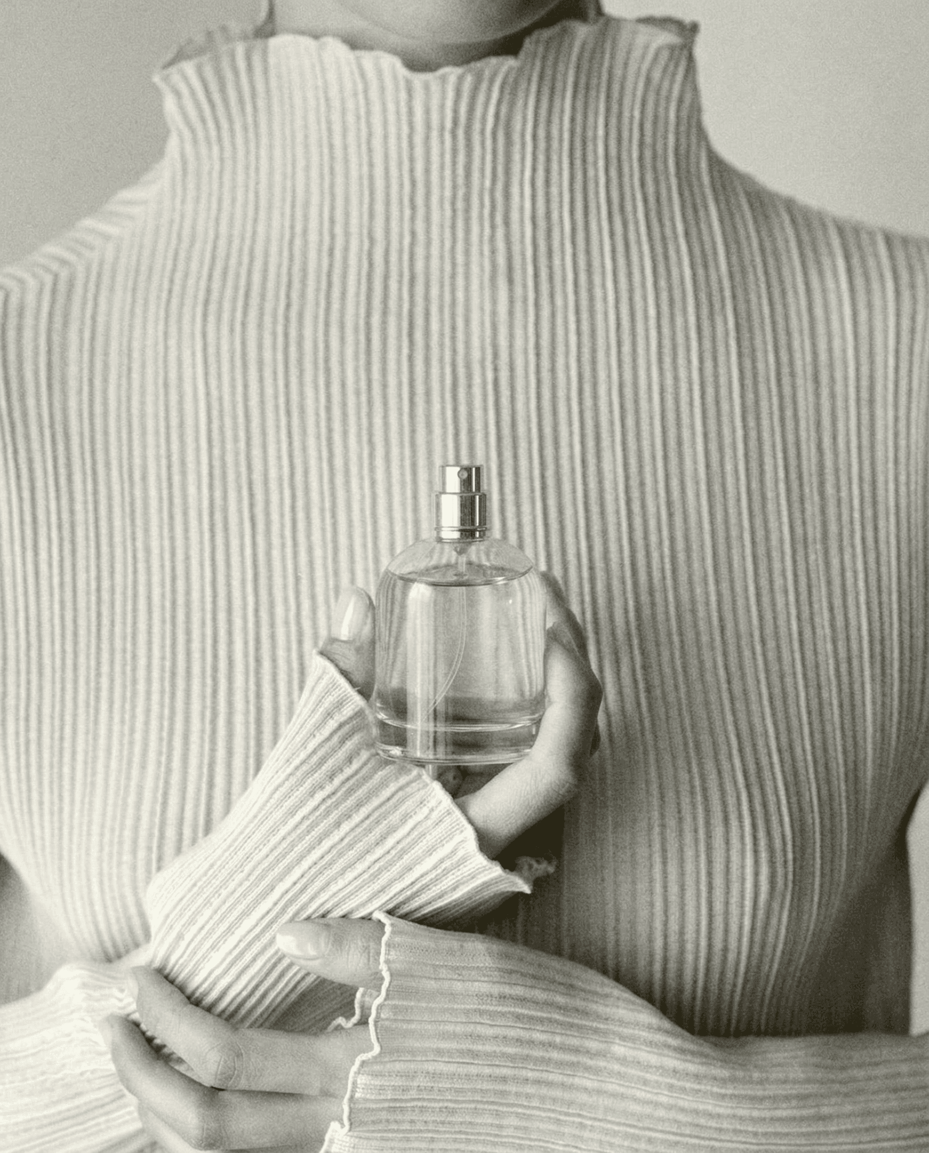 Cropped intimate torso shot of a figure in a high-necked ribbed pleated garment holding a minimalist clear glass fragrance bottle pressed against the chest at heart level with both hands. Symmetrical centered composition. Soft diffused black and white film photography aesthetic with gentle grain. The vertical pleated fabric lines frame the bottle. Quiet luxury, artisanal niche fragrance mood. Nostalgic, tender, poetic. No face visible.