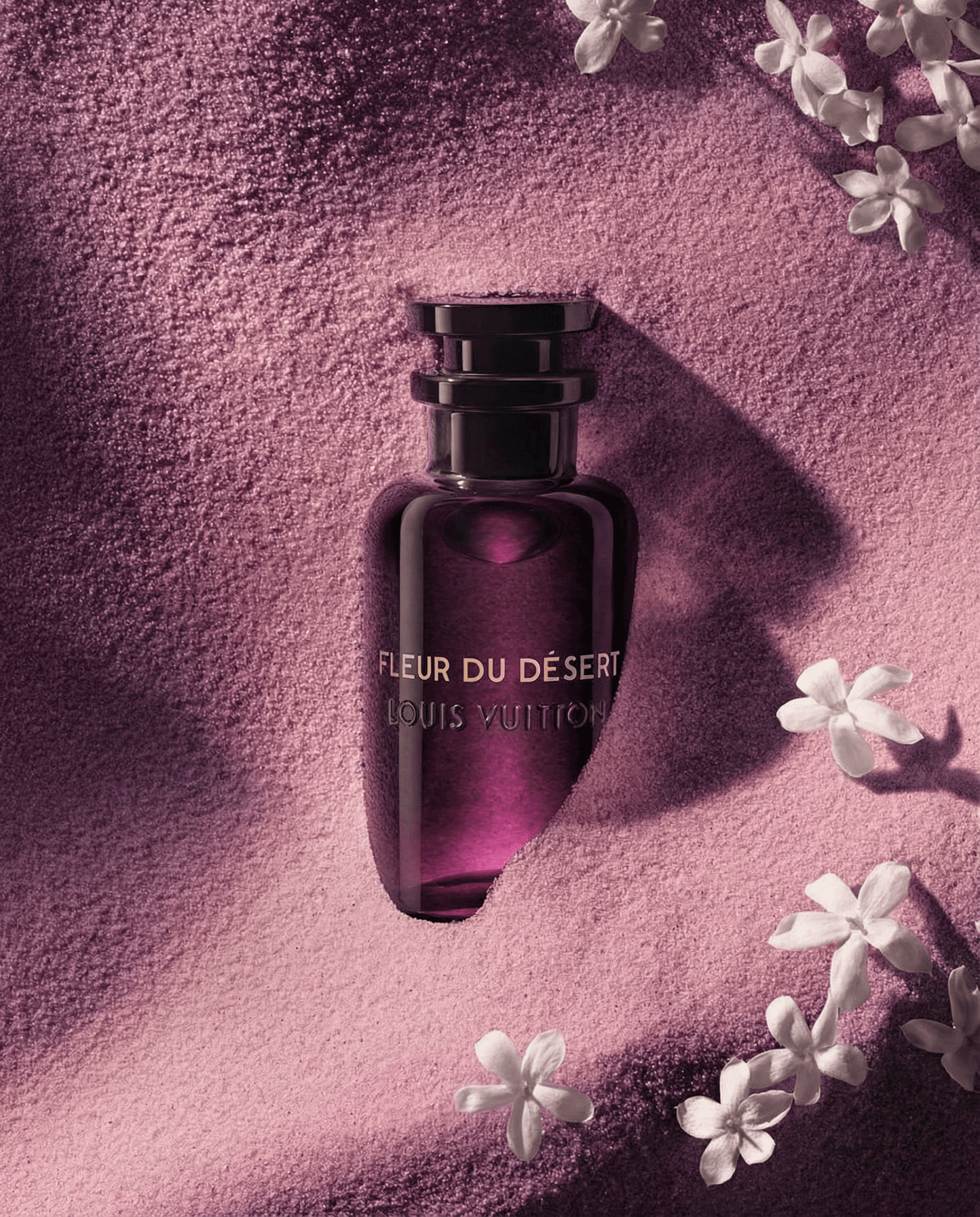Flat-lay product shot of a deep amethyst glass perfume bottle on fine lavender-pink desert sand, delicate white jasmine blossoms scattered elegantly in the corners, soft overhead diffused lighting with gentle shadows, monochromatic mauve palette with the bottle catching light to reveal an inner gradient from dark plum to bright magenta-violet, serene and opulent luxury fragrance aesthetic
