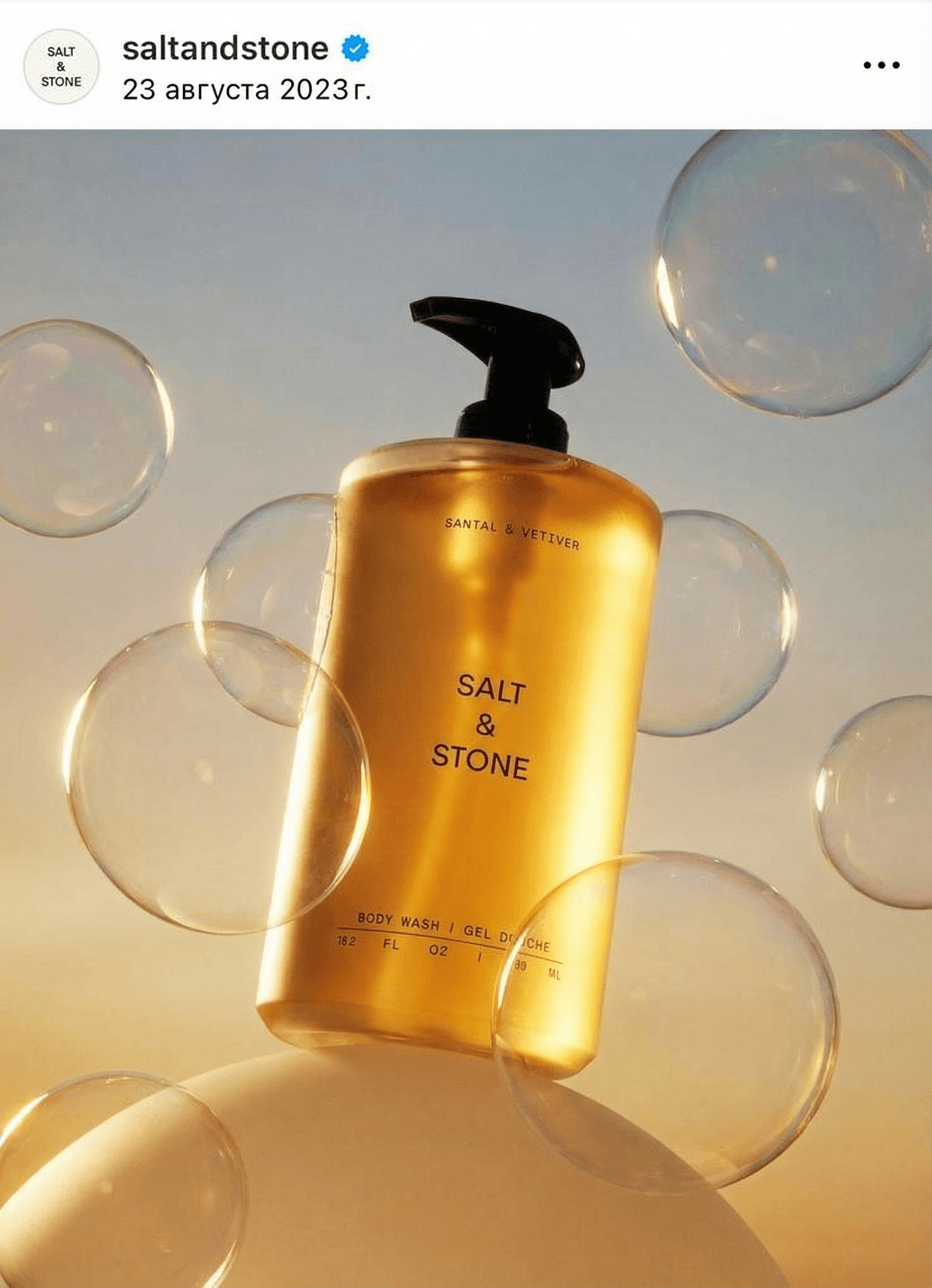 The product sits on a smooth rounded sculptural cream-colored prop, several large perfectly formed translucent iridescent soap bubbles floating around it, warm golden back-lighting illuminating the product from behind creating a luminous halo effect, dreamy gradient background blending pale blue-gray with warm golden tones, the liquid inside the bottle glows amber in backlight, golden hour atmosphere, Californian sun-drenched luxury aesthetic, warm and aspirational, professional product photography