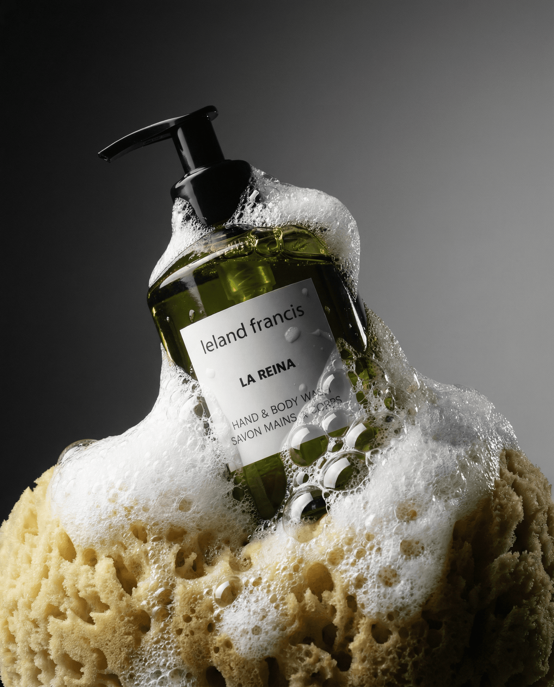The product is nestled into a natural sea sponge surrounded by rich white sudsy foam and lather, dramatic moody studio photography, seamless gradient background transitioning from dark charcoal gray on the left to medium gray on the right, strong directional key light from upper left creating deep shadows, specular highlights on the packaging, wet foam bubbles catching light, luxury spa aesthetic, editorial high-contrast lighting, dark and sophisticated, 4K product photography