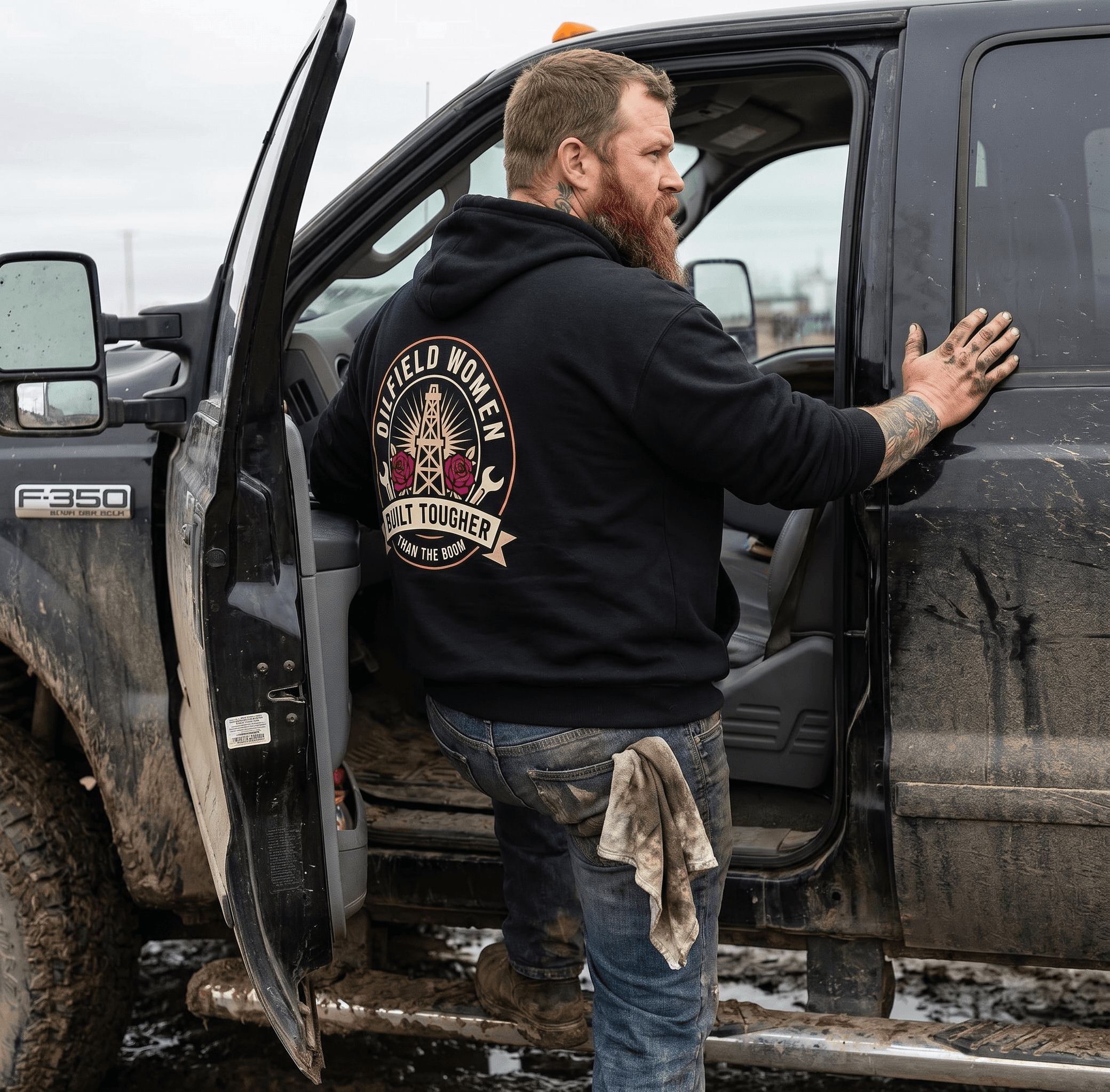 A man in his early 40s with a full reddish-brown beard, faded neck tattoo visible above the hoodie collar, full forearm sleeve tattoos visible at the wrists, broad shoulders, grease-stained calloused hands, leaning back against the open driver-side door of a muddy black Ford F-350 work truck in a prairie service yard. Wearing this exact "Forged in Mud Apparel" hoodie (preserve the design exactly — black hoodie with the large white circular FMA monogram badge centered on the back). Photographed from a three-quarter back angle so the badge reads clearly and large. Overcast Alberta daylight, dried mud on the truck doors, an oily rag tucked into his back jeans pocket, scuffed steel-toe boots. Authentic worksite photography — not posed, no smile, looks like a candid shot a coworker took on lunch break.