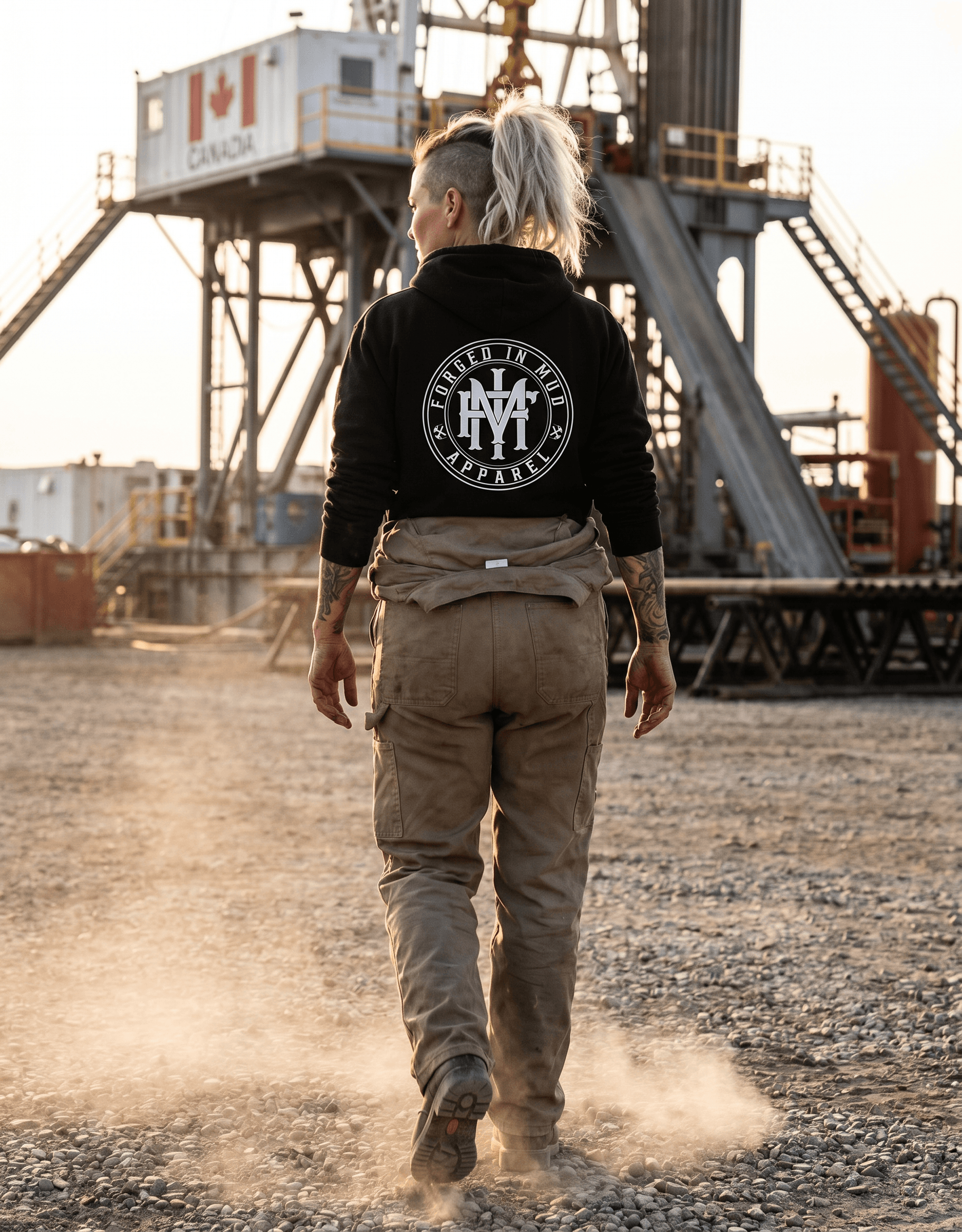 A woman in her late 30s with a half-shaved head and a messy bleached ponytail, full sleeve tattoos on both arms partially visible at the wrists, weathered tanned hands, wearing this exact "Oilfield Women Built Tougher" hoodie (preserve the design and colors exactly — black hoodie with the circular badge featuring a derrick, pink roses, wrenches and cream banner) under an unzipped FR coverall pulled down around her waist with the sleeves tied at the hips. She is walking across a gravel pad toward a Canadian drilling rig at golden hour, photographed from directly behind so the back design reads clearly and large. Steel-toe boots, dust kicked up at her ankles, slightly underexposed iPhone photo quality, candid documentary feel — looks shot on a coworker's phone, not a campaign. Real worker body proportions, not a model.