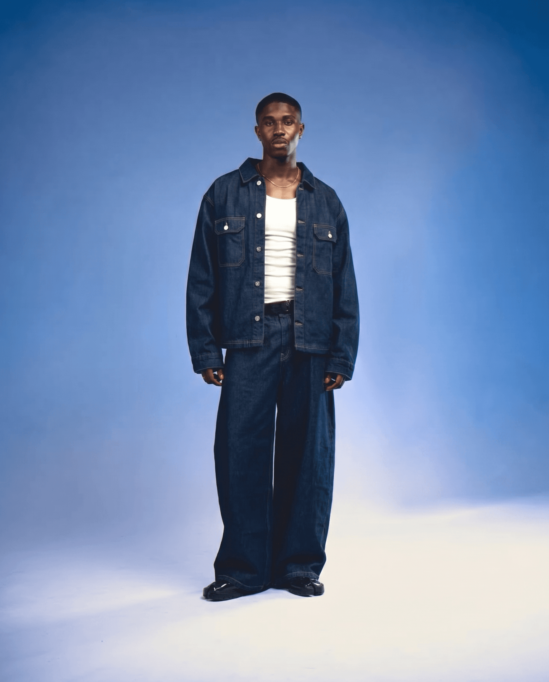 A tall male model stands in a seamless studio with a gradient backdrop transitioning from periwinkle cornflower blue to white at the floor, wearing indigo denim-on-denim — an oversized raw denim trucker jacket unbuttoned over a white ribbed tank, paired with extremely wide-leg dark denim jeans and black polished shoes, thin gold chain necklace, soft atmospheric haze on the studio floor, controlled diffused studio lighting with a gentle glow, deep indigo, white, and blue gradient palette, high-fashion contemporary menswear editorial, 4K