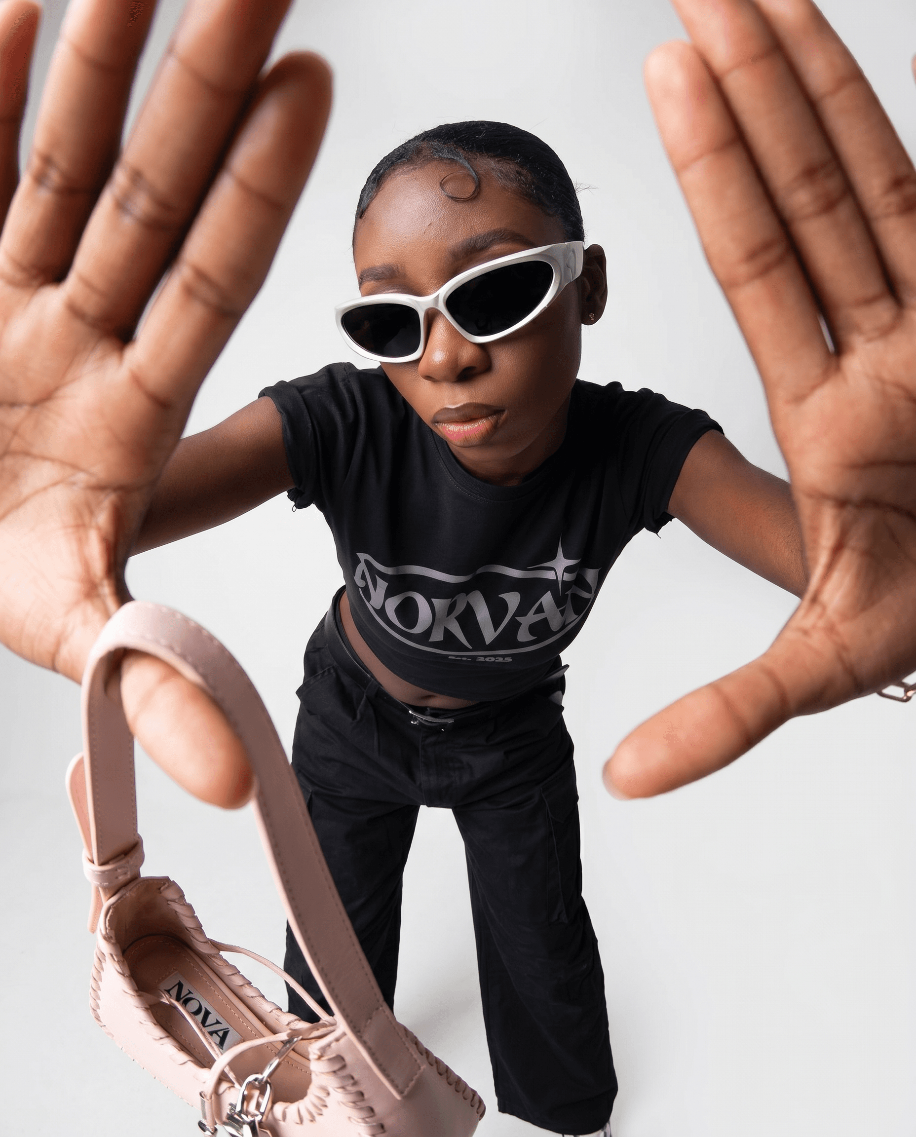 A female model with slicked-back cornrows reaches boldly toward the camera in an extreme close-up fisheye-distorted wide angle, wearing a black graphic cropped baby tee and black cargo wide-leg trousers, white futuristic cat-eye wrap sunglasses, holding a blush pink strappy sandal as a prop, bright high-key studio lighting against a clean white-grey seamless backdrop, palette of black, white, blush pink, and rich skin tones, confrontational Gen-Z streetwear brand campaign, bold and disruptive, 4K