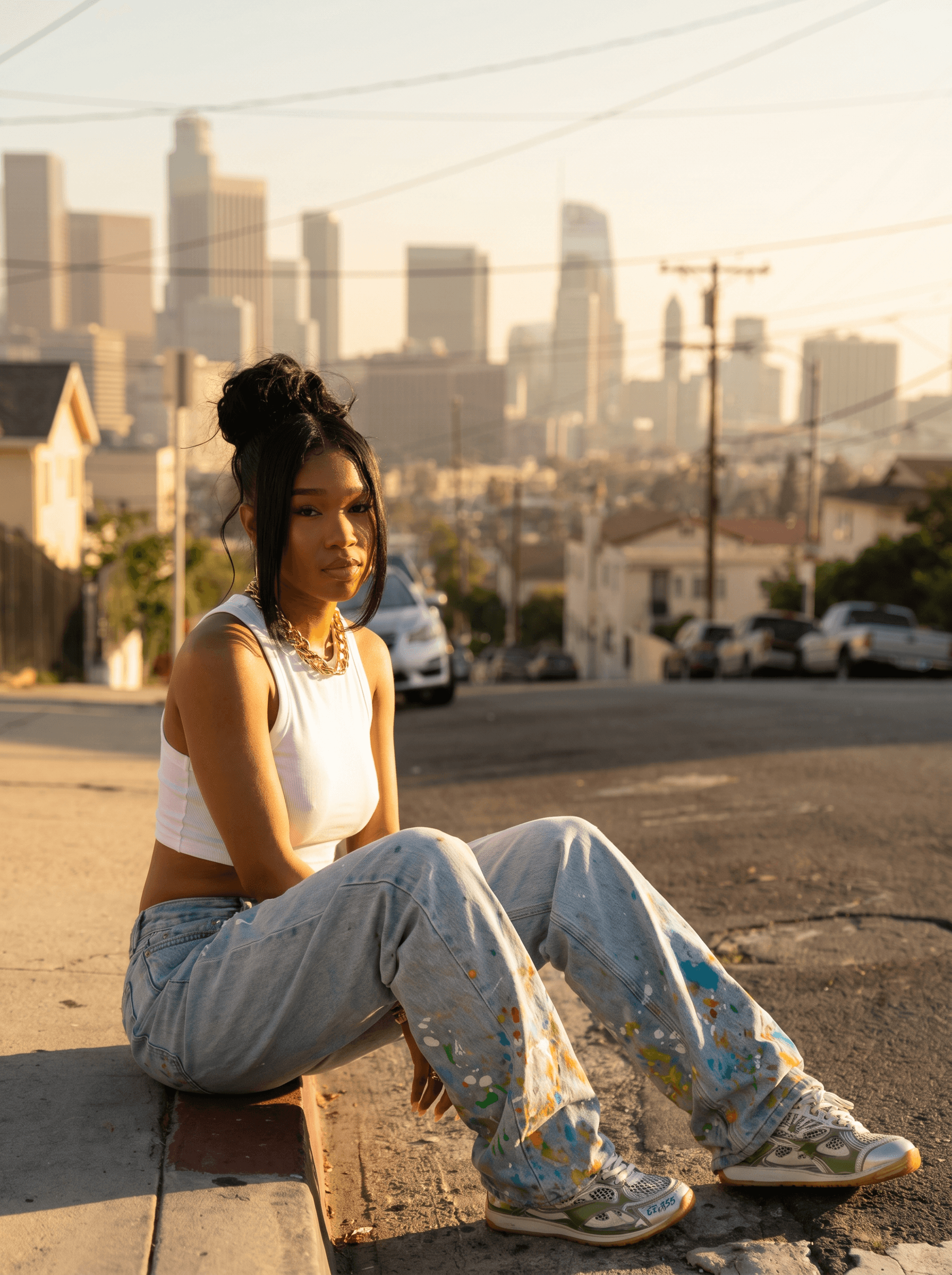 A female model with a high messy bun sits on a residential LA street curb, downtown skyline hazy in the background, wearing a white cropped tank, paint-splattered light-wash baggy jeans and chunky sneakers, layered gold chunky chain necklace and bracelets, warm golden-hour sunlight casting sun-drenched glow on the skin with hazy atmospheric cityscape behind, color palette of white, light denim blue, gold, and sky blue, California golden-hour street style editorial, 4K