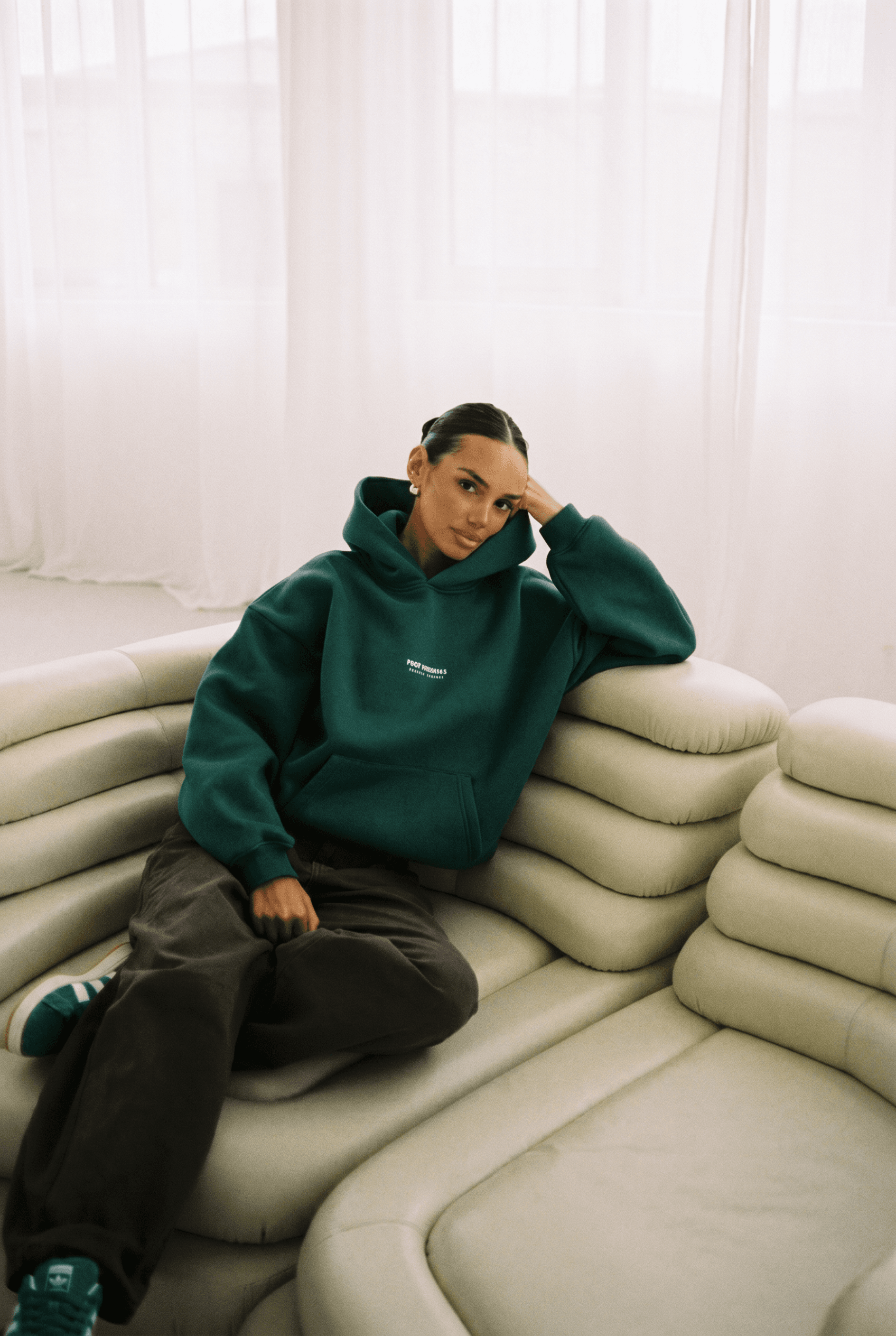 A female model with a sleek tight low bun and small gold hoop earrings is seated on a sculptural cream modular cloud-like sofa, wearing a deep forest-green oversized hoodie and dark charcoal wide-leg trousers with green-and-white Adidas sneakers, sheer white curtains diffusing soft warm natural light in the background, cozy low-contrast analog film warmth, palette of forest green, cream, ivory, charcoal brown, and white, elevated quiet-luxury loungewear Scandinavian-streetwear editorial, 4K