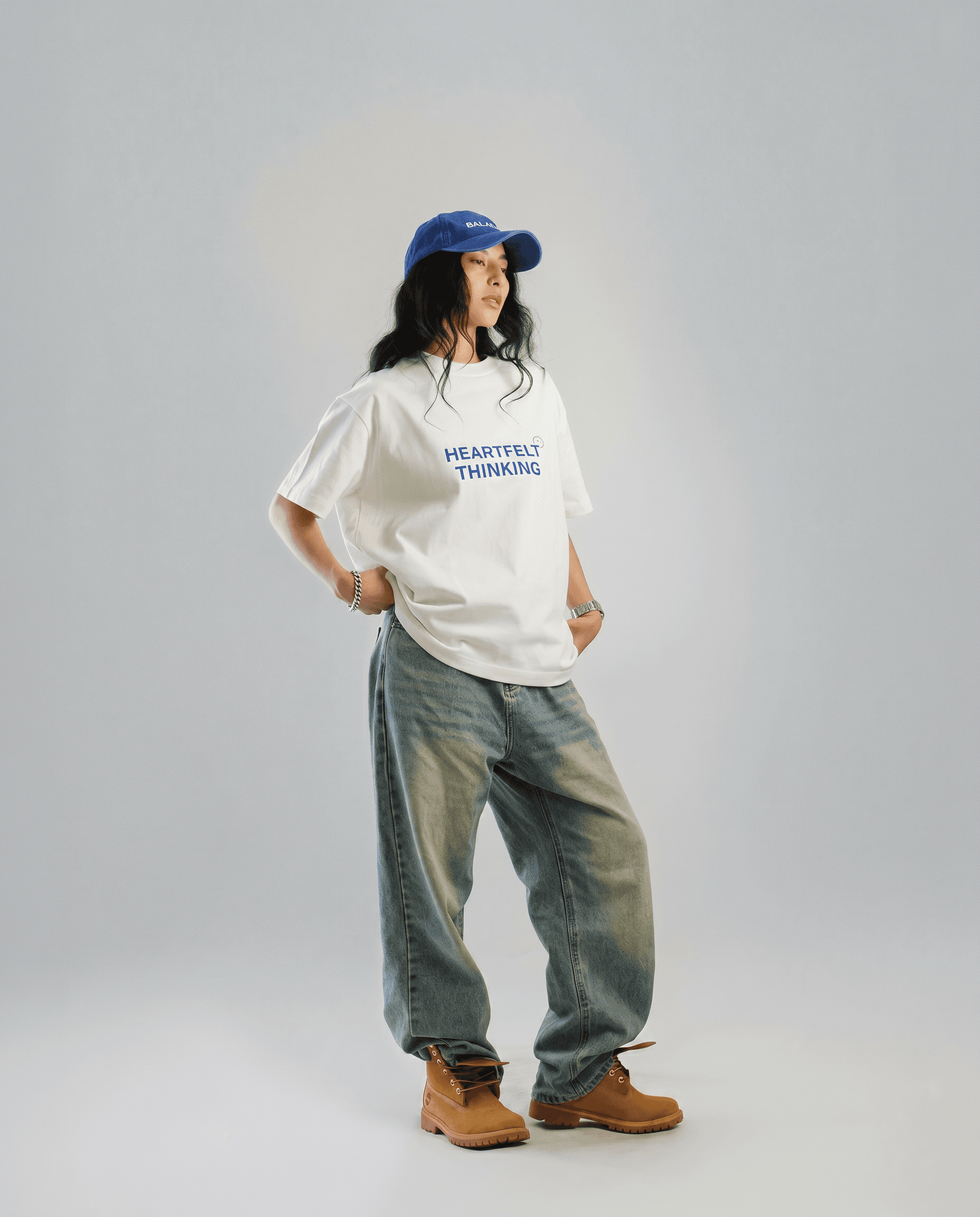 A female model with long wavy black hair tucked under a royal blue dad cap stands against a clean neutral grey studio seamless backdrop, wearing an oversized white graphic t-shirt, very baggy faded light-wash jeans with a worn vintage look and wheat tan Timberland-style boots, silver bracelet, bright even commercial studio lighting with minimal shadows, palette of white, royal cobalt blue, faded denim, wheat tan, and grey, Y2K early-2000s hip-hop nostalgia streetwear lookbook, 4K