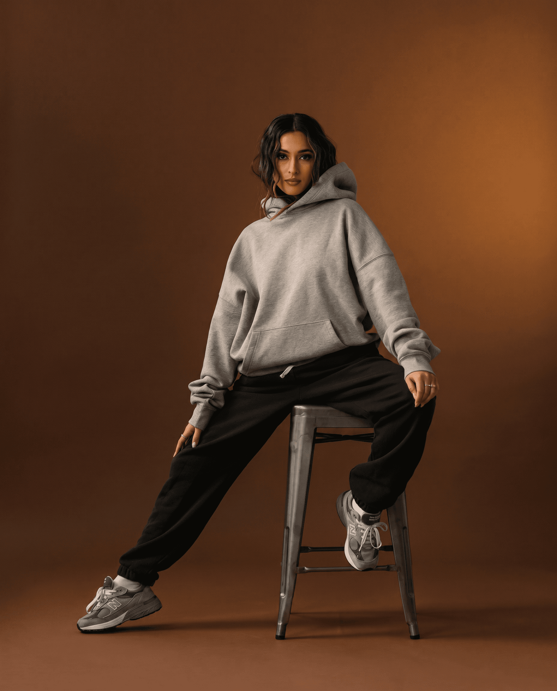 A female model with long dark wavy hair sits casually on a metal industrial stool against a warm chocolate brown caramel seamless studio backdrop, wearing an oversized heather gray hoodie and black oversized sweatpants, chunky gray New Balance dad sneakers, minimal jewelry, moody warm studio lighting with intimate quality creating a golden glow on the warm backdrop contrasting with the cooler gray outfit tones, heather gray, charcoal black, warm chocolate brown, and silver-gray palette, premium athleisure elevated streetwear Essentials Fear of God editorial, 4K