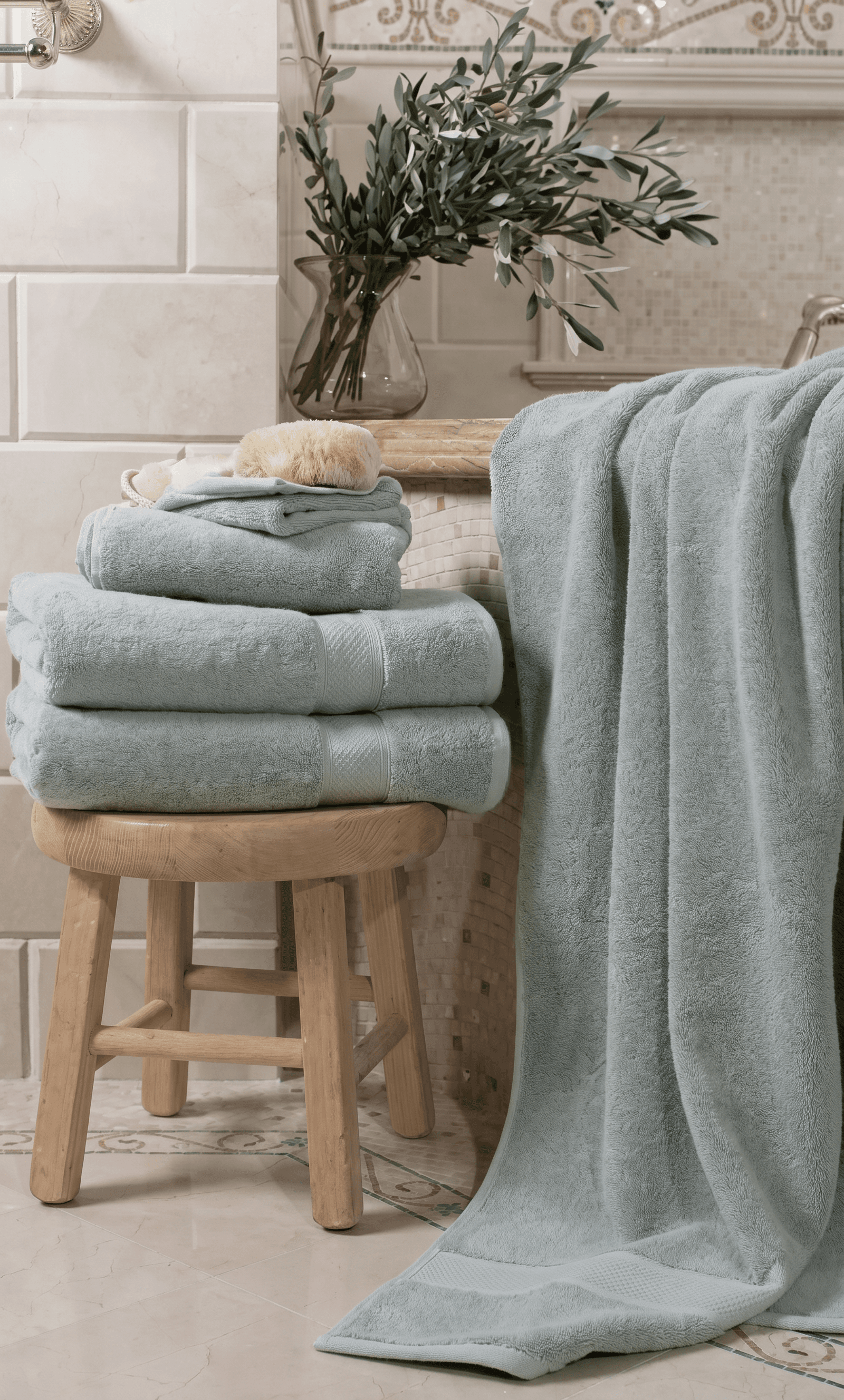 Mediterranean spa bathroom editorial, plush duck-egg blue cotton towels neatly stacked on a rustic light-wood stool, one draped over a bathtub edge, cream subway tiles with decorative border, olive branch in glass vase, natural bristle brush, tranquil and luxurious mood