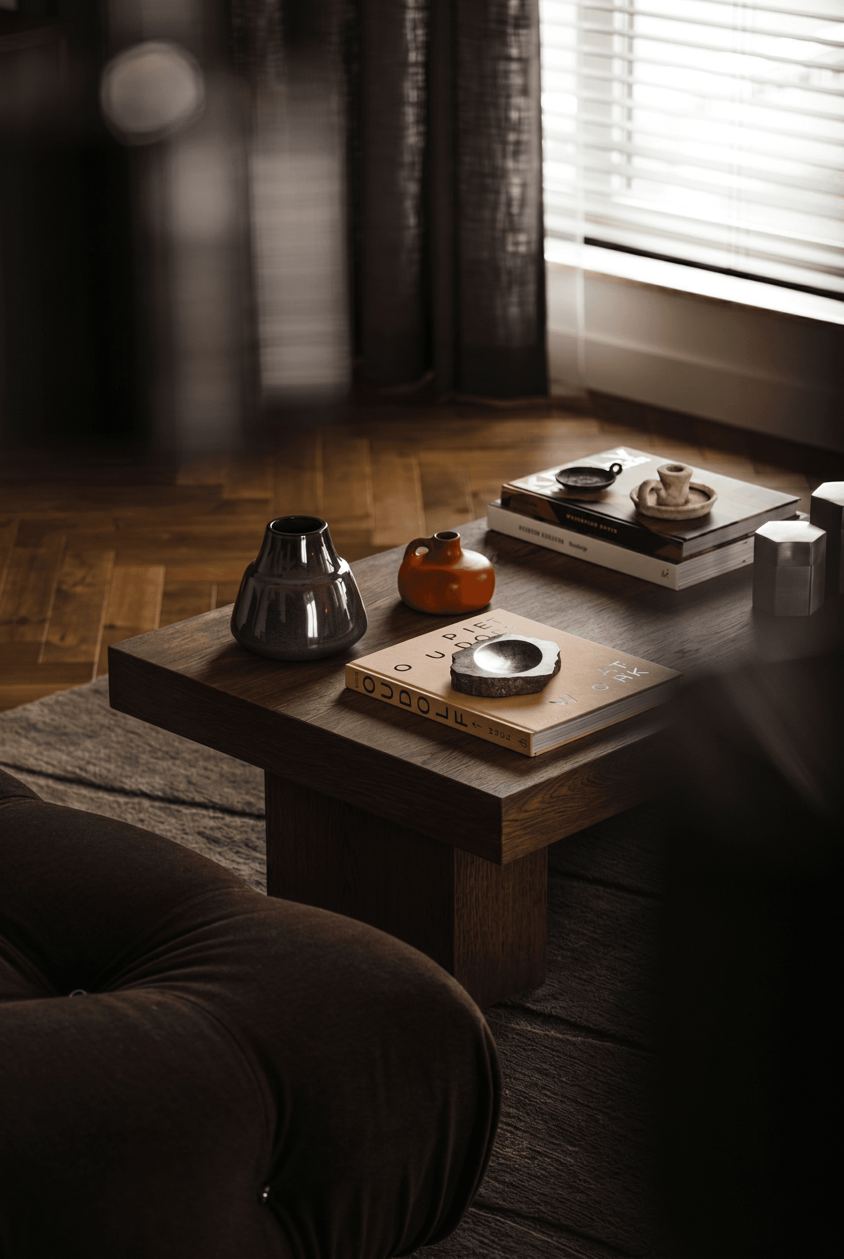 Masculine collector interior, dark walnut modernist coffee table styled with smoky grey glass vase, burnt-orange ceramic vessel, stacked art books and stone dish, moody dimly-lit living room with herringbone parquet, dark curtains, venetian blinds filtering soft light