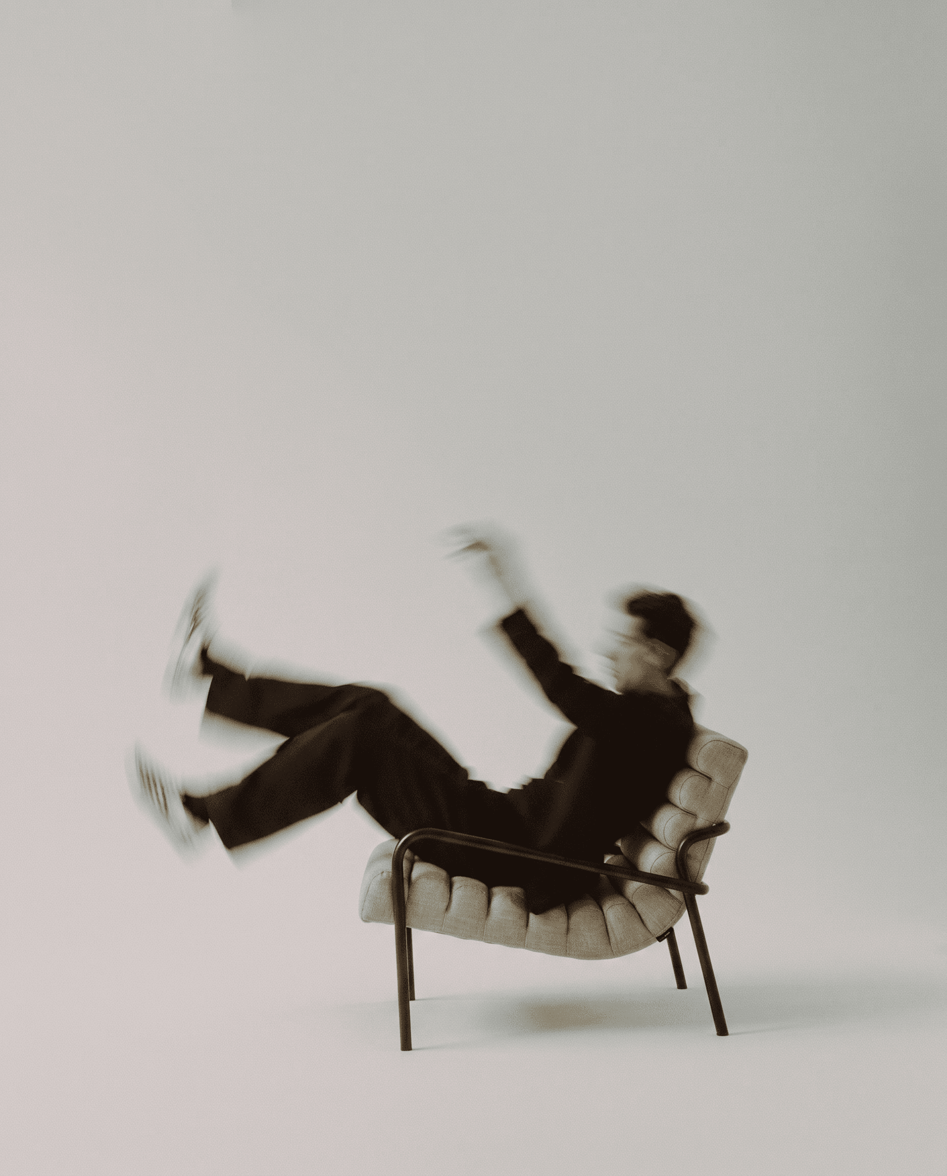 Fashion-forward editorial, slim dark metal frame lounge chair with oatmeal channel-tufted cushioning, motion-blurred figure in all black falling backward into the chair, minimal pale grey studio backdrop, dynamic avant-garde energy blending furniture design with human movement