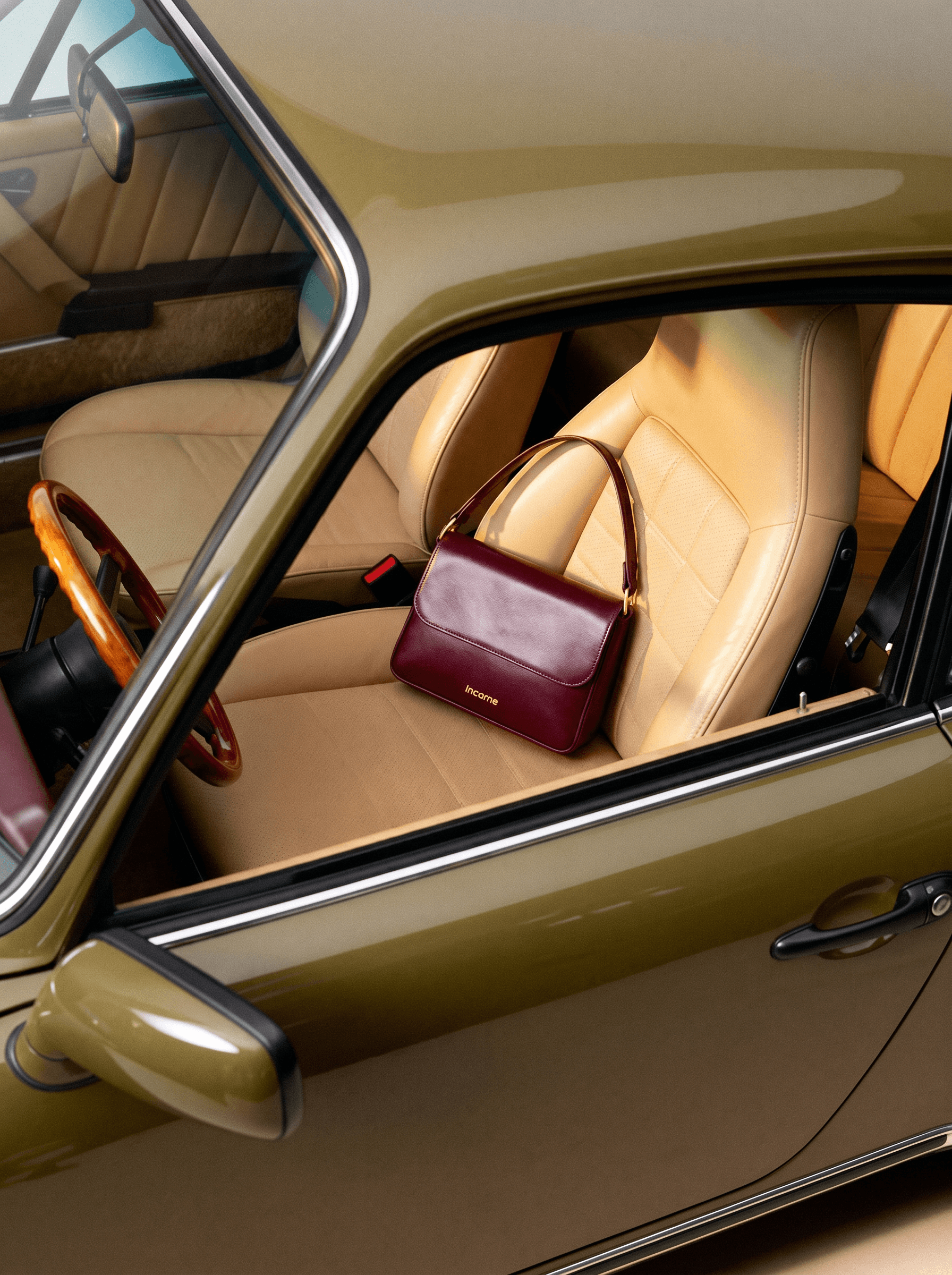 Retro-cinematic luxury editorial, compact structured oxblood leather shoulder bag with gold hardware resting on a tan quilted leather seat of a vintage olive-green sports car, wood-rimmed steering wheel visible, European glamour and timeless cinematic style