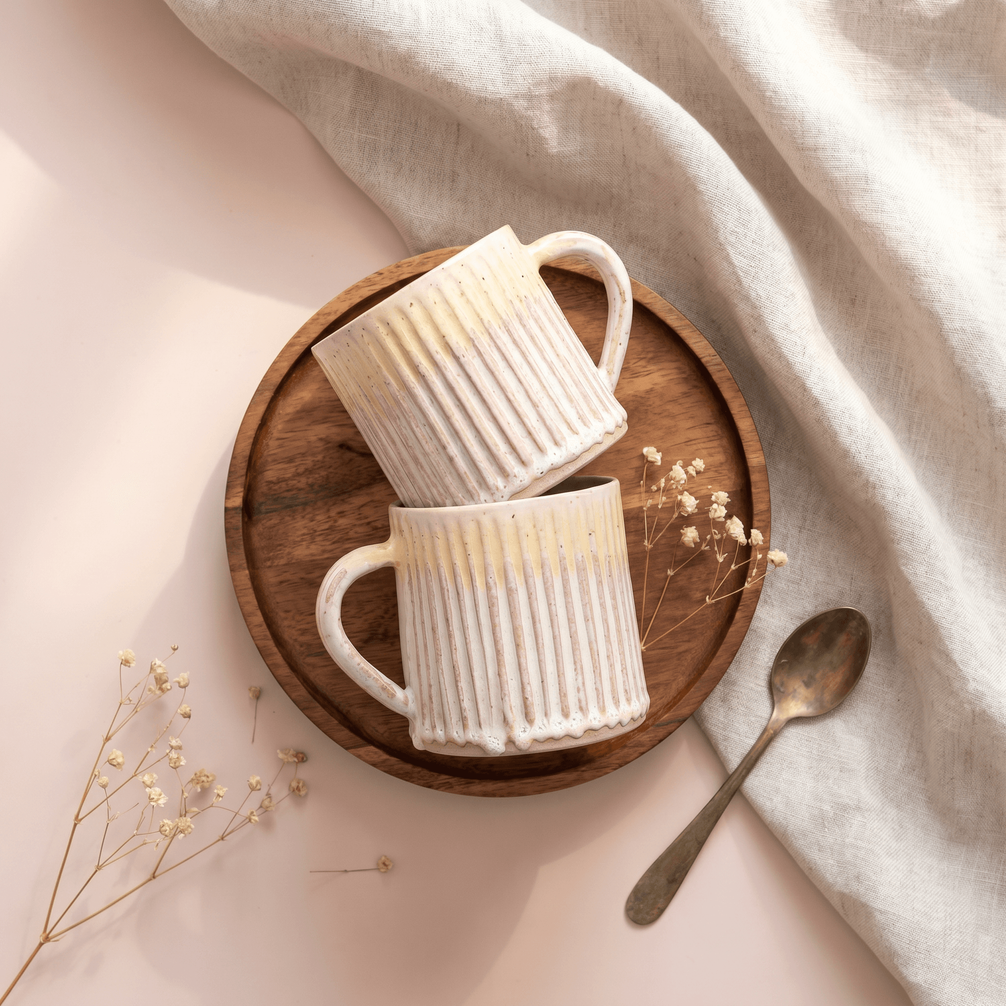 Artisanal lifestyle flat-lay, two handcrafted vertically ribbed ceramic mugs in creamy white with speckled glaze stacked on a round acacia wood tray, delicate dried baby's breath flowers and brass spoon, soft blush backdrop with draped linen fabric, warmly organic and handmade