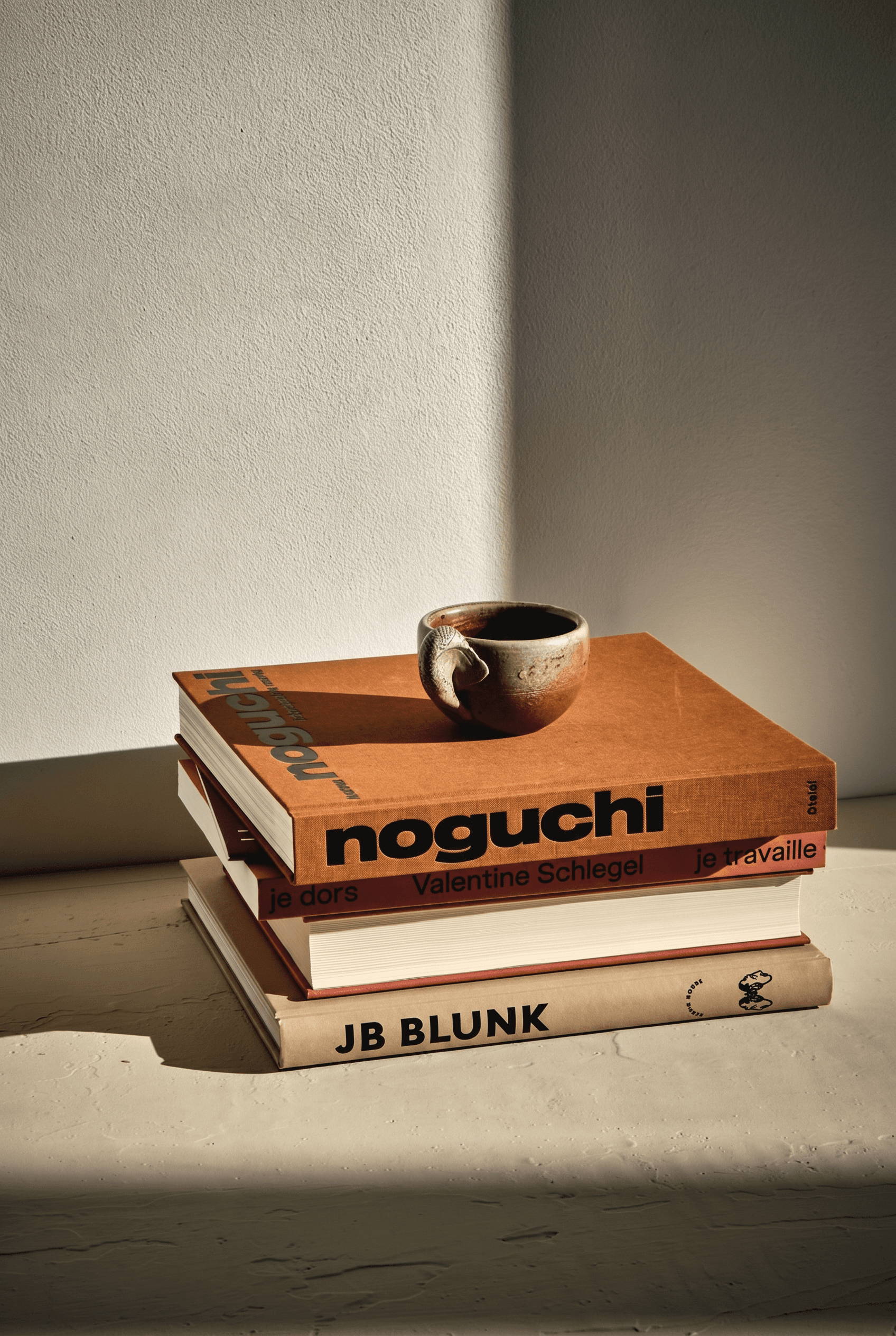 Intellectual design-conscious flat-lay, curated stack of art coffee table books in burnt orange and coral tones topped with a handmade earth-glazed ceramic cup, light stone surface with dramatic diagonal natural light casting deep shadows, warmly minimal contemplative mood