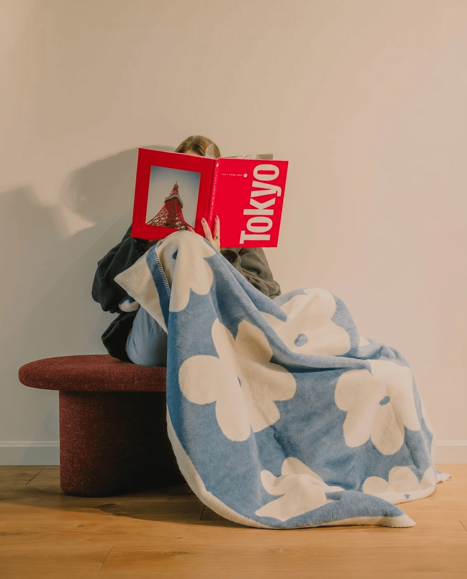 Contemporary home lifestyle, sky-blue knit blanket with large white abstract floral motifs draped over a person on a burgundy bouclé ottoman, reading a bold red coffee table book, white wall with warm wood flooring, playful design-forward and casual-chic mood