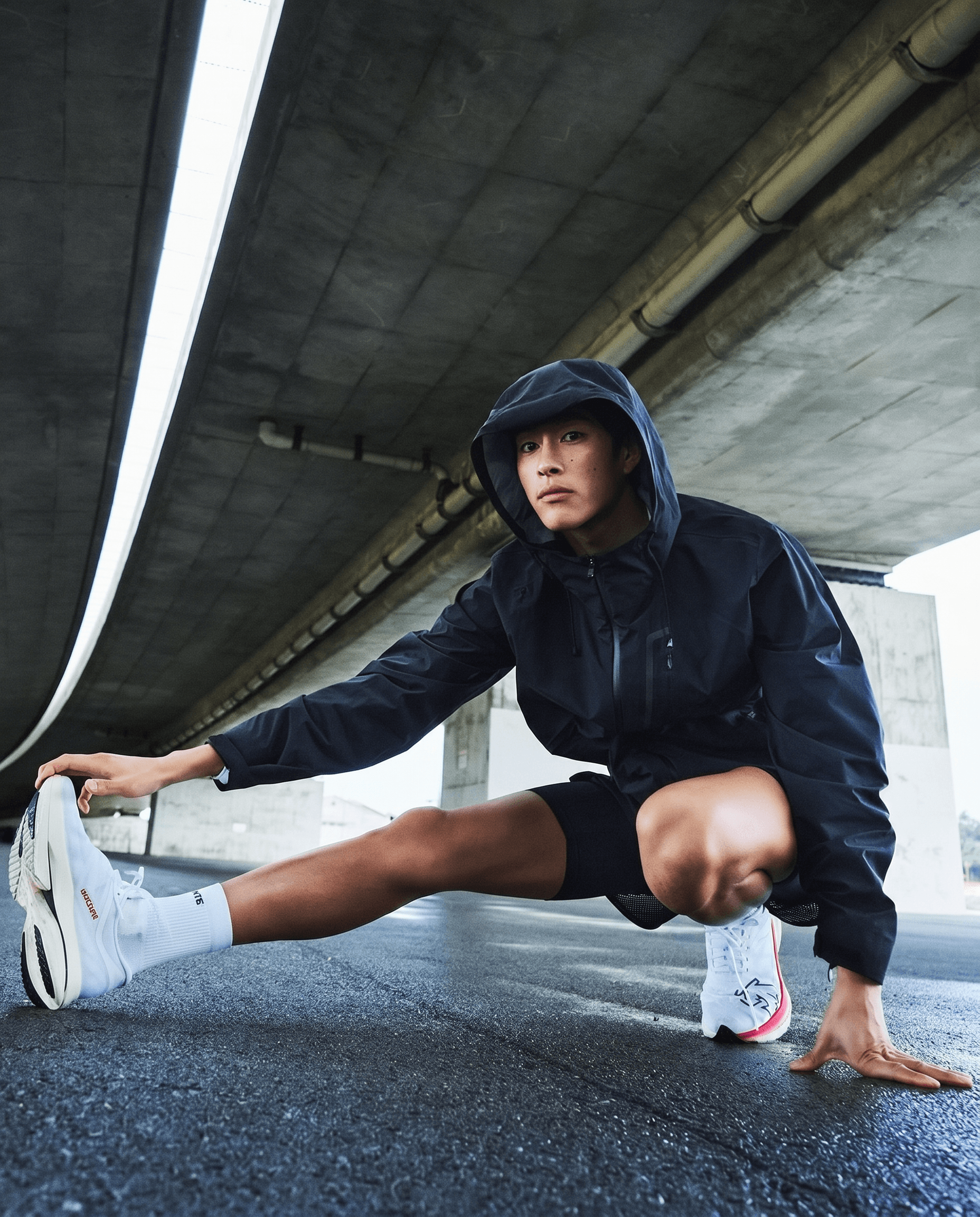 Gritty urban athlete editorial, dark navy performance hooded windbreaker and black shorts with white running shoes, model in a deep lateral lunge under a raw concrete overpass on wet glistening asphalt, moody grey tones with dramatic low angle framing, determined intense urban runner campaign energy