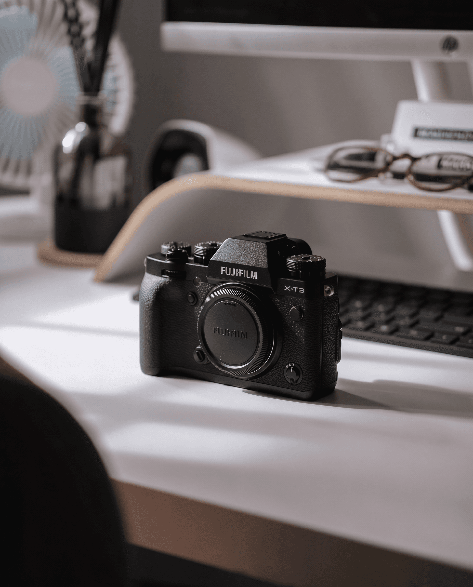 Authentic lifestyle still life, Fujifilm X-T3 mirrorless camera body resting on a clean white desk with soft daylight filtering through nearby windows, shallow depth of field with warm bokeh from a monitor, keyboard and desk accessories blurred in the background, muted blacks and clean whites, calm minimalist work-from-home aesthetic with intimate natural lighting