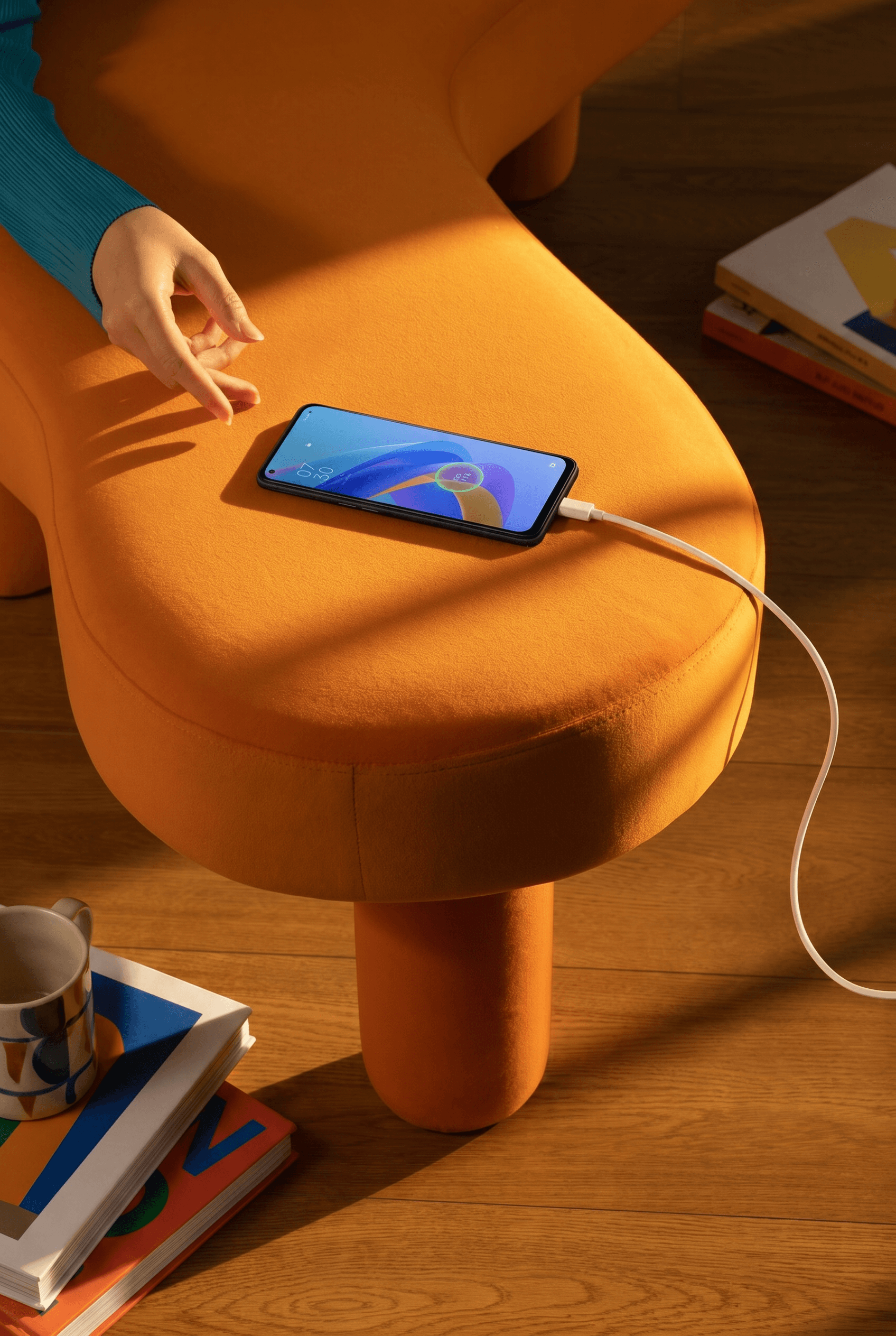 Warm playful interior lifestyle moment, smartphone charging on a bold mustard-orange velvet ottoman with chunky sculptural rounded legs, vibrant color pop of teal sleeve entering frame, warm honey-toned wooden floor planks below, the rich contrast between the tangerine orange upholstery and cool teal tones creating a joyful contemporary design-conscious atmosphere with abundant natural light