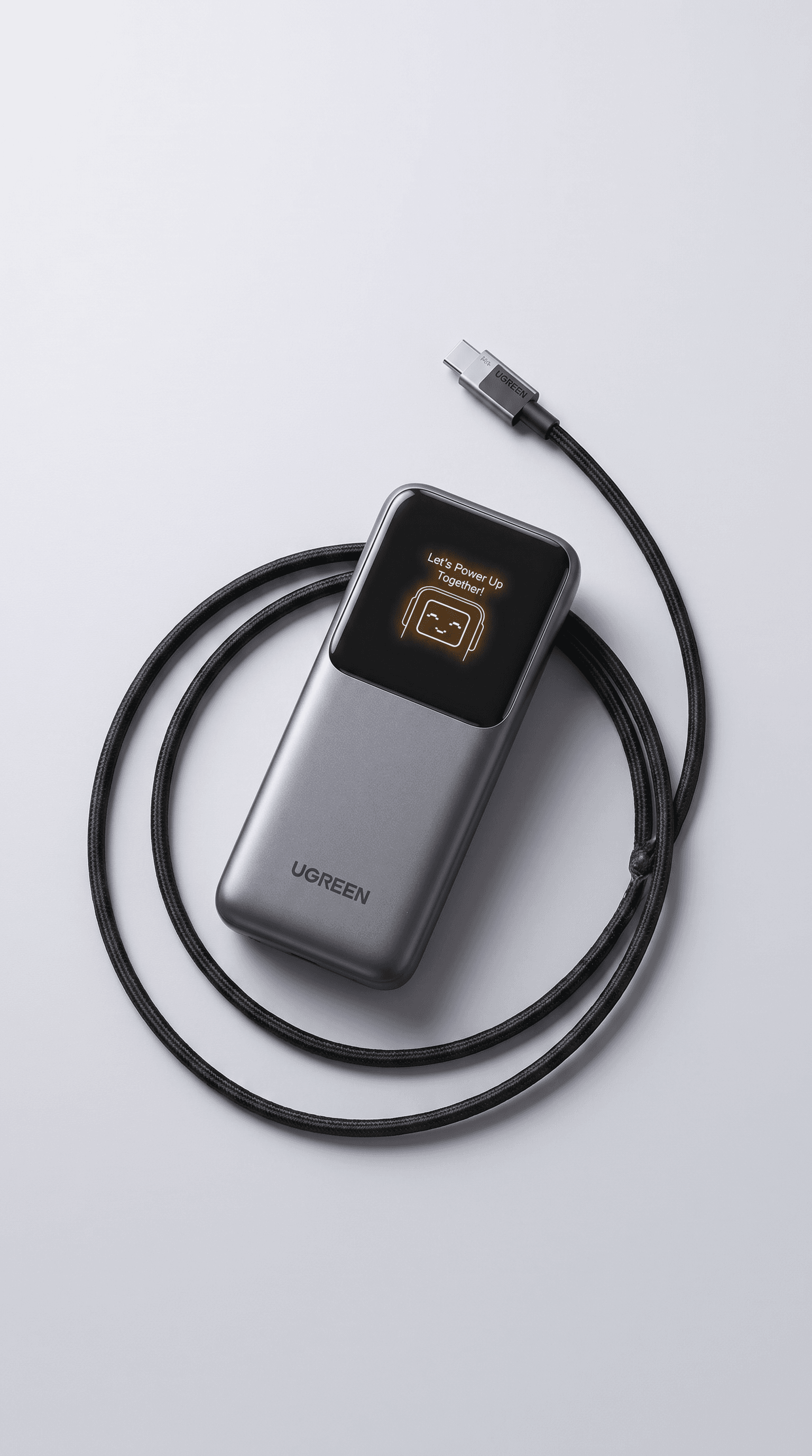 Clean playful tech flat-lay, gunmetal gray UGREEN portable power bank on a smooth light gray surface with a black braided USB-C cable artfully coiled around it, the small OLED display showing a cute robot mascot face glowing warmly, soft diffused overhead studio lighting with subtle shadows, the minimalist composition balancing utilitarian precision with personality and charm