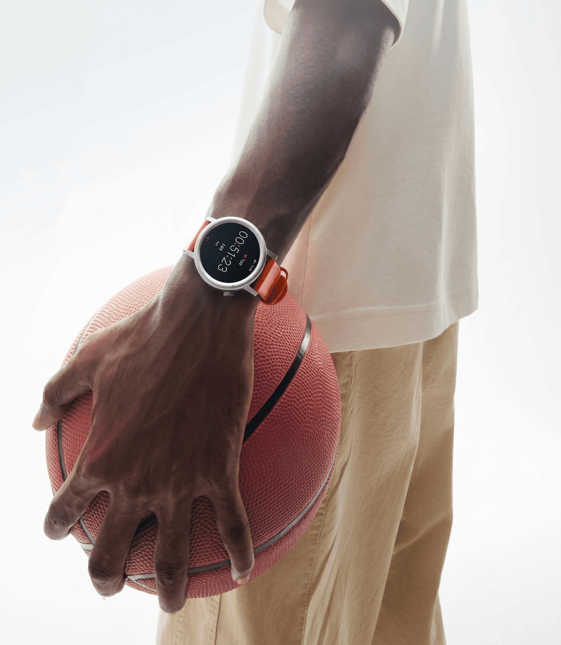 Athletic lifestyle studio portrait, round-face smartwatch with bold orange sport band on the wrist of a person holding a textured basketball at their side, clean white studio background with soft wrap-around lighting, tan khakis and white tee, the stopwatch display reinforcing athletic performance, fresh energetic sports editorial mixing casual cool with everyday functionality