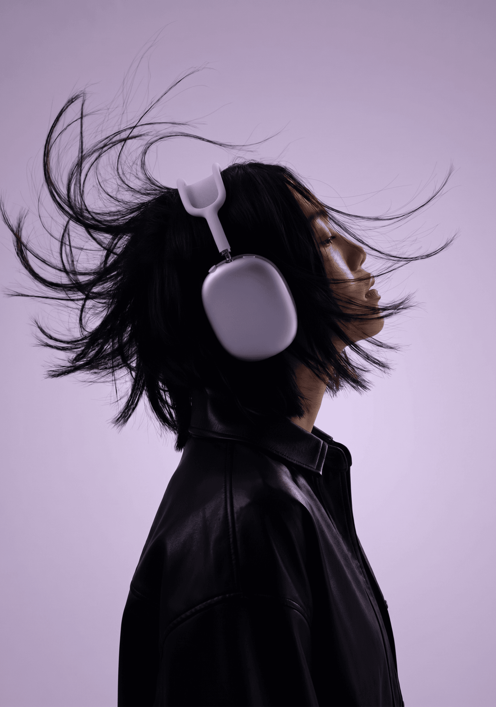 High-energy fashion editorial, woman in black leather jacket wearing lavender Apple AirPods Max, wind machine sending dark hair flying dramatically, monochromatic soft purple-lavender background perfectly matching the headphones color, profile composition with strong directional light, the kinetic movement of hair conveying sonic power and immersive listening, bold fashion-tech crossover energy with chromatic cohesion