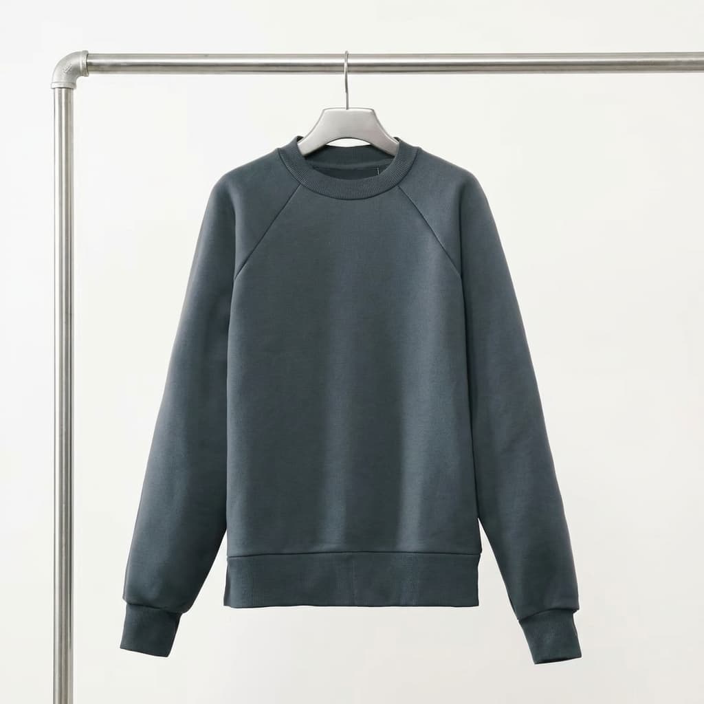 The photo shows a minimalist product shot of a crewneck sweatshirt hanging neatly on a hanger.
The garment is centered in the frame, suspended from a clean, industrial-style metal clothing rail. The sweatshirt has a relaxed, classic silhouette with raglan sleeves, ribbed cuffs, hem, and neckline. Its fabric appears smooth and structured, suggesting a mid- to heavyweight knit designed to hold its shape.
The color is a muted charcoal or blue-gray tone, understated and versatile. There is no visible branding on the front, reinforcing a quiet, utilitarian aesthetic.
The background is pure white and evenly lit, creating strong contrast and isolating the product completely. The composition is symmetrical and uncluttered, drawing attention to the form, construction, and texture of the garment.
Overall, the image feels clean, functional, and modern, typical of premium e-commerce or lookbook photography where clarity and material quality are the focus.