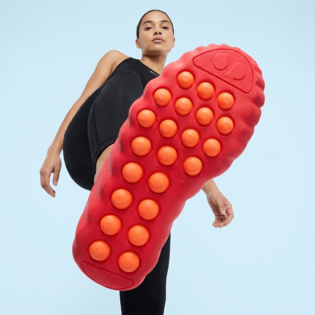 The photo is a dynamic, fashion-forward action portrait shot from an extreme low angle.
A model strides forward with one foot lifted directly toward the camera, making the sole of the shoe dominate the frame. The oversized, textured outsole—bright red with raised circular tread elements—becomes the primary visual focus, appearing exaggerated and powerful due to perspective.
The body is partially foreshortened, with the torso and head receding into the background. The pose feels assertive and in motion, as if caught mid-step. The model’s expression is neutral and composed, reinforcing confidence rather than aggression.
Lighting is bright and even, giving the image a crisp, high-energy look. The pale, almost sky-like background removes any environmental context, allowing the graphic shapes and bold colors of the footwear to stand out.
Overall, the image feels bold, playful, and modern, using scale distortion and perspective to emphasize design, movement, and attitude—typical of contemporary fashion or footwear campaigns that want impact and visual immediacy.