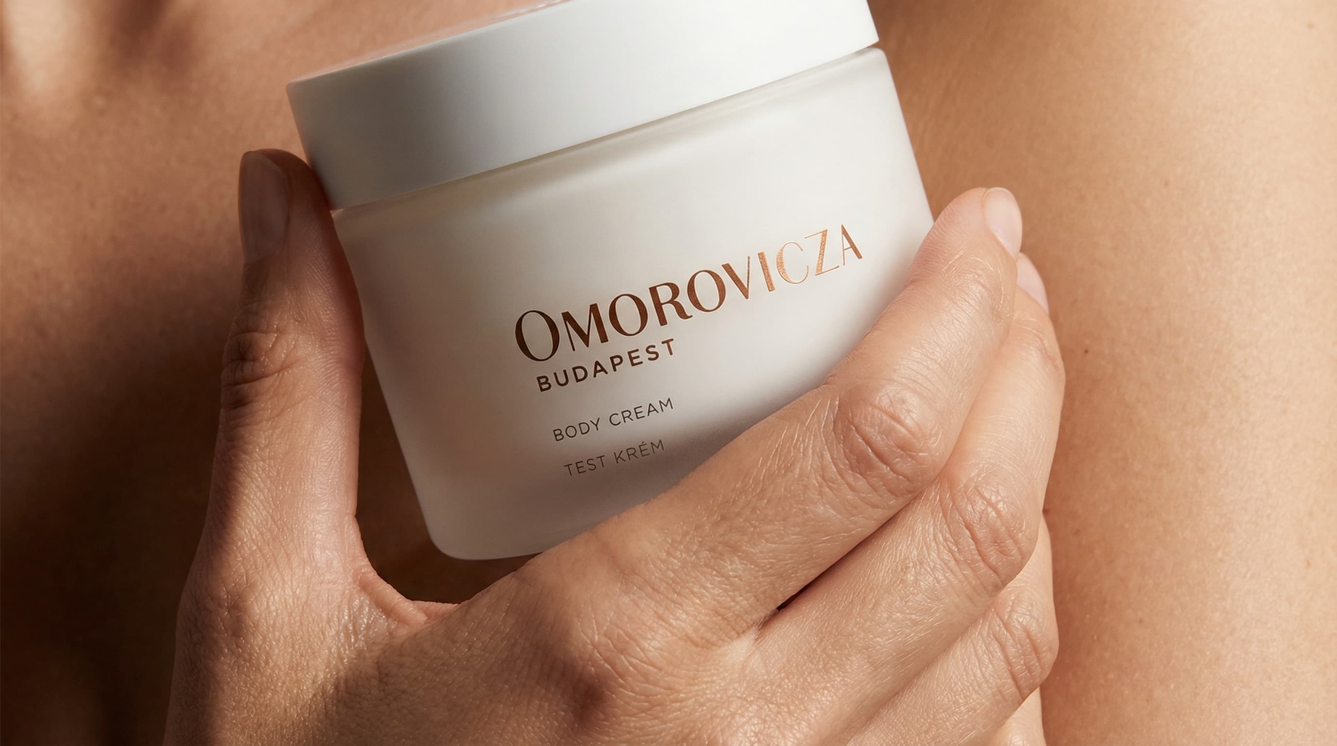 The photo is a refined, intimate body-care product portrait with a strong emphasis on skin and simplicity.
A jar of body butter is held gently in one hand, positioned against bare skin that fills the background. The close framing removes any environmental context, making skin tone, texture, and product design the primary visual elements.
The packaging is minimal and neutral, with clean typography and a soft, matte finish that reinforces a modern, dermatology-inspired aesthetic. The hand placement feels natural and supportive rather than posed, suggesting ease and everyday use.
Lighting is soft and directional, creating subtle highlights and shadows that accentuate the contours of the body and the smooth surface of the container. Skin texture is visible and natural, contributing to a sense of authenticity and tactility.
Overall, the image feels calm, modern, and sensorial, balancing clinical credibility with warmth and intimacy—well suited to contemporary skincare branding focused on trust and real results.