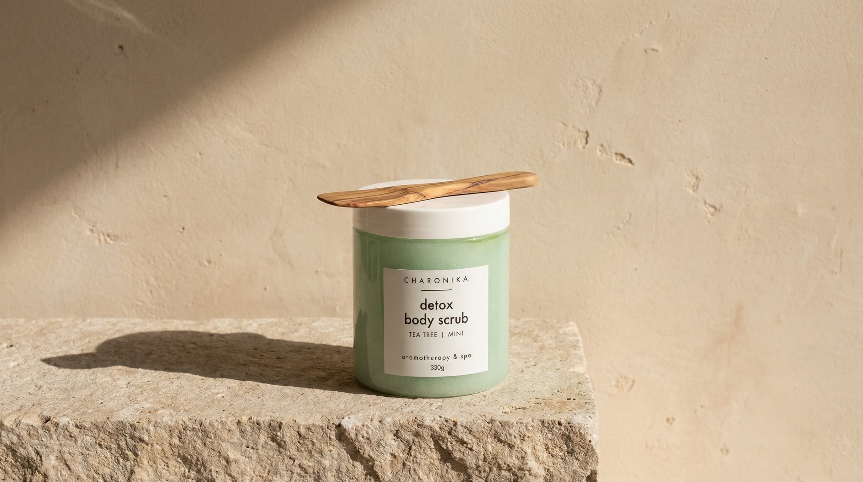 The photo is a quiet, sculptural product still life with a natural, editorial sensibility.
A minimal, cylindrical skincare jar sits centered on a rough stone surface, presented like a small object of ritual. A wooden spatula rests casually on top of the lid, adding a tactile, handmade detail that suggests mindful use.
The background is a warm, textured wall with subtle imperfections, giving the scene an architectural, almost Mediterranean feel. Natural light enters from the side, casting a soft shadow that cuts across the surface and wall, creating depth and a sense of time passing.
The color palette is restrained—soft beige, stone, and warm neutrals—allowing material textures to take precedence over color. The composition is calm and balanced, with plenty of negative space around the product.
Overall, the image feels earthy, slow, and considered, blending skincare with lifestyle and atmosphere rather than overt product display.
