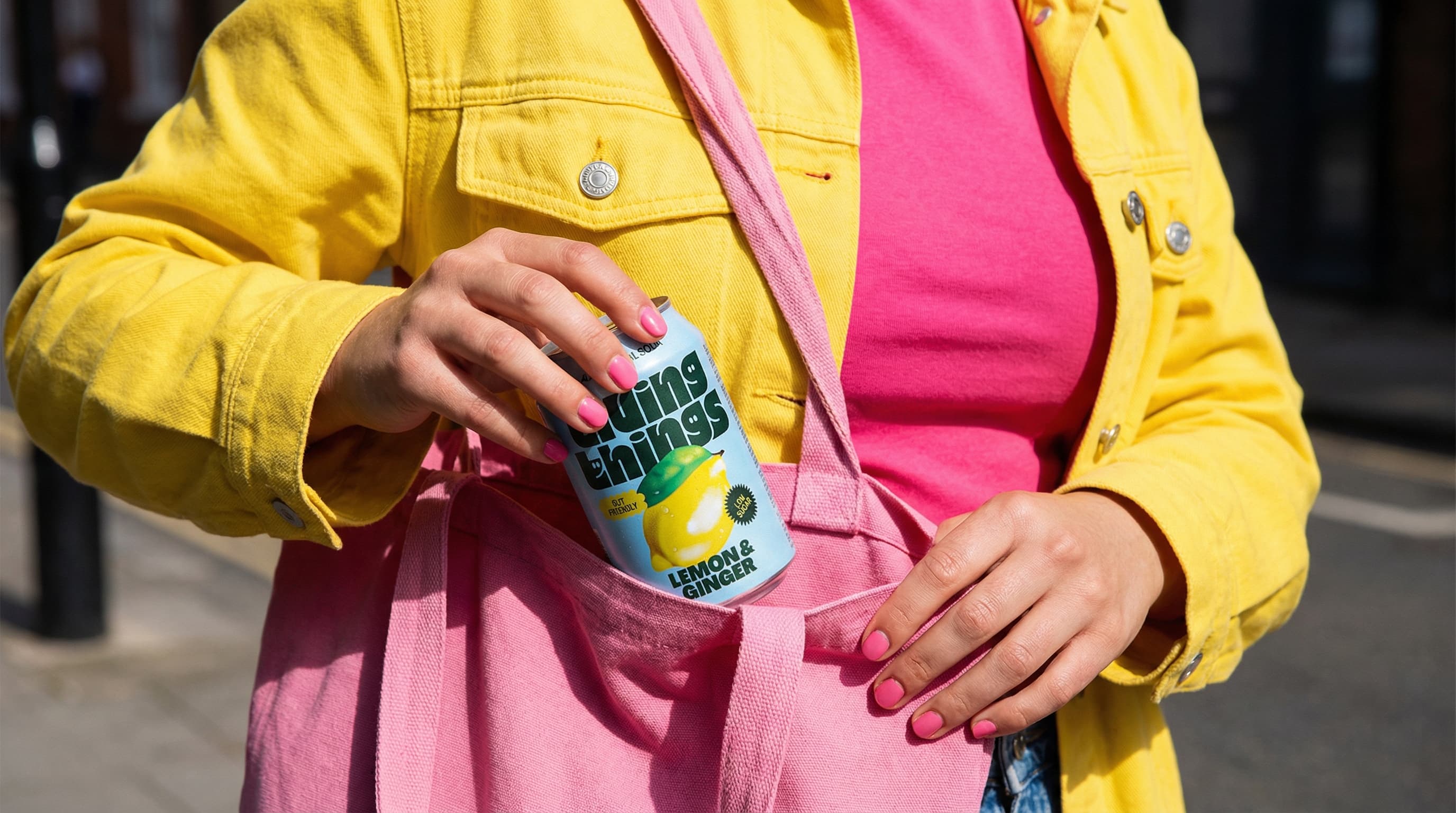 The image is a bold, lifestyle product shot driven by color and everyday motion.
A cropped view of a person’s torso and hands fills the frame. They’re dressed in bright, saturated layers—a yellow jacket over a pink top—with a pink canvas tote slung over the shoulder. One hand grips the tote strap while the other holds a tall beverage can halfway inside the bag, as if mid-movement.
The can’s graphic design stands out sharply against the clothing, becoming the focal point through contrast rather than isolation. Matching pink nail polish subtly echoes the palette, tying the styling together in a deliberate but casual way.
Lighting is direct and punchy, creating crisp shadows and enhancing the vibrancy of the colors. The framing feels candid and in-motion, like a moment captured while heading out, rather than a staged studio still.
Overall, the image feels energetic, youthful, and lifestyle-driven, positioning the product as part of an active, colorful, on-the-go routine rather than a hero object on display.