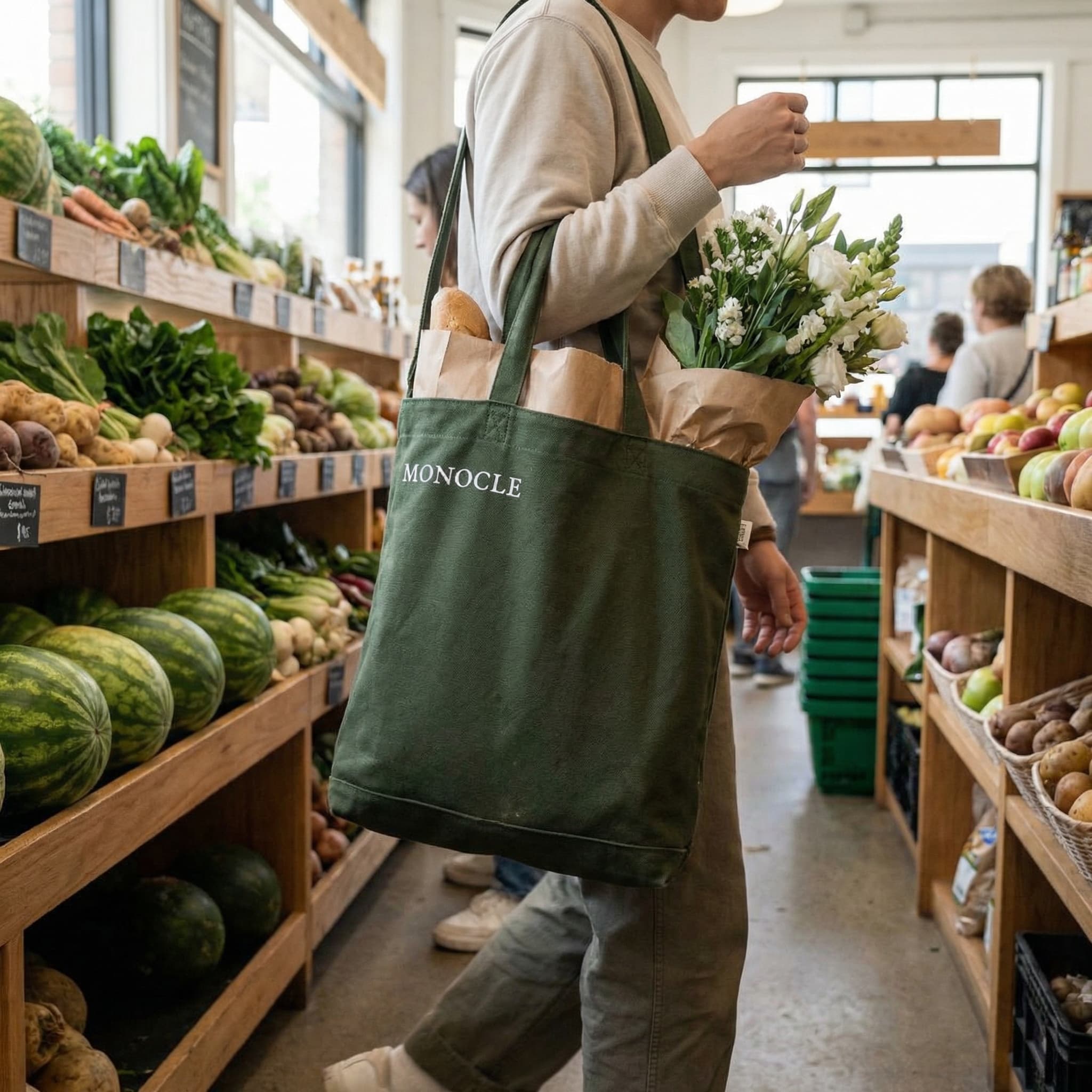 The photo captures a grounded, everyday lifestyle moment rooted in routine and place.
A person stands in front of a produce display, holding a dark green canvas tote filled with groceries and a bouquet of white flowers. The tote hangs naturally at their side, slightly worn and heavy, suggesting use rather than styling. Paper grocery bags peek out from the top, reinforcing the sense of a real errand mid-flow.
Behind them, wooden shelves are stacked with fresh produce—watermelons, leafy greens, and vegetables—creating a dense, textured backdrop. The scene feels candid and observational, as if caught while moving through the store rather than posed.
The color palette is earthy and muted: greens, browns, and soft neutrals dominate, giving the image a calm, organic tone. Lighting appears natural and ambient, consistent with an indoor market environment.
Overall, the image feels casual, local, and lifestyle-driven, positioning the branded tote as a quiet marker of identity and habit rather than a focal product. It communicates values—freshness, routine, taste—through context and atmosphere.