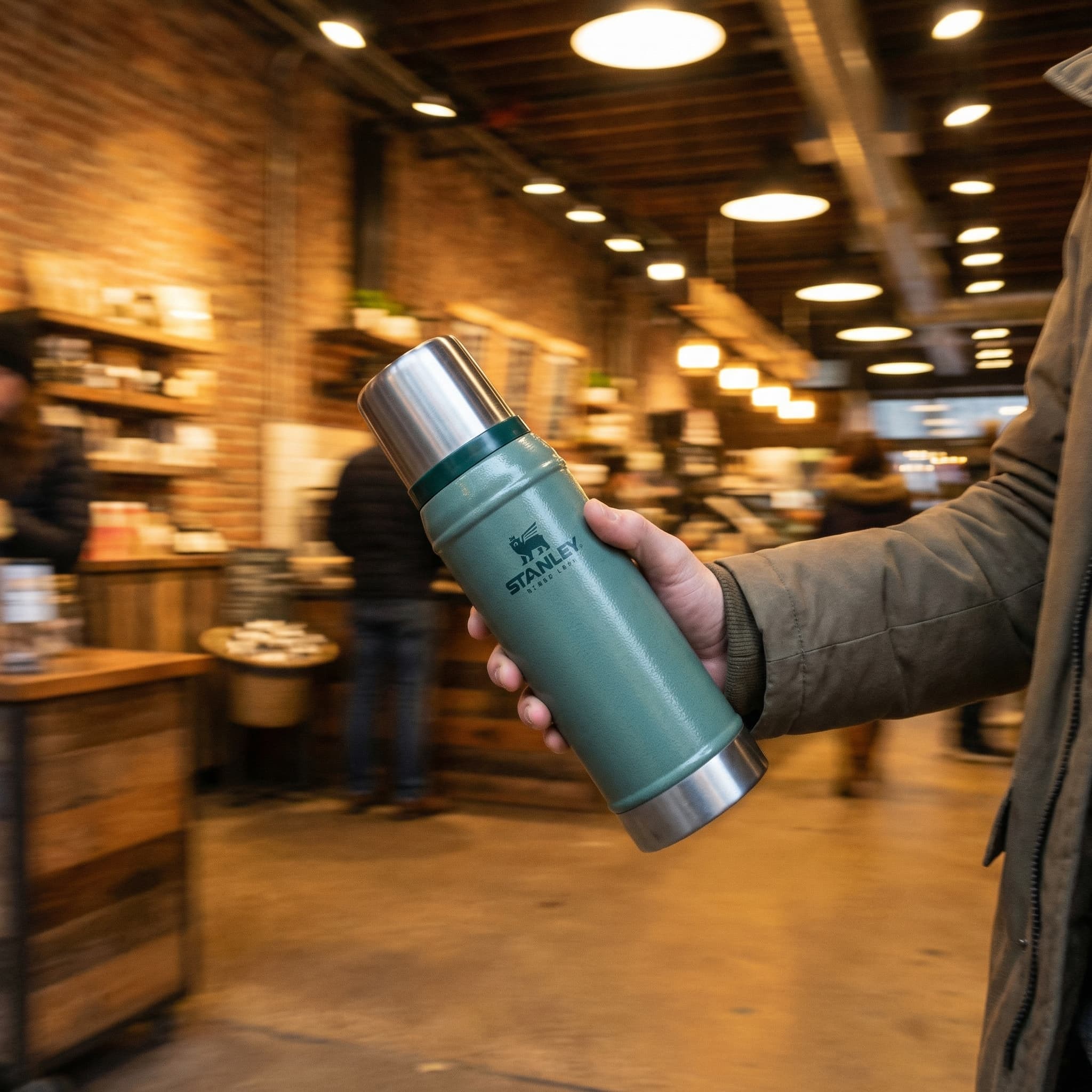 The image captures a moody, lifestyle-driven product moment with an emphasis on movement and atmosphere.
A hand carries a large insulated tumbler mid-stride, angled slightly forward as if walking through an indoor space. The product is clearly present but not centered or isolated—its presence feels incidental, part of real life rather than staged display.
The background is heavily motion-blurred, with warm lights and brick textures streaking horizontally, suggesting movement through a café or urban interior. This blur contrasts with the relative sharpness of the hand and tumbler, reinforcing the feeling of momentum and everyday use.
Lighting is warm and low, creating a cozy, evening ambiance. The muted teal of the tumbler stands out against the golden and brown tones of the environment, giving the product visual clarity without breaking the natural mood.
Overall, the image feels casual, kinetic, and lived-in, positioning the product as a companion in motion—something carried through daily routines rather than showcased in a static, polished setting.